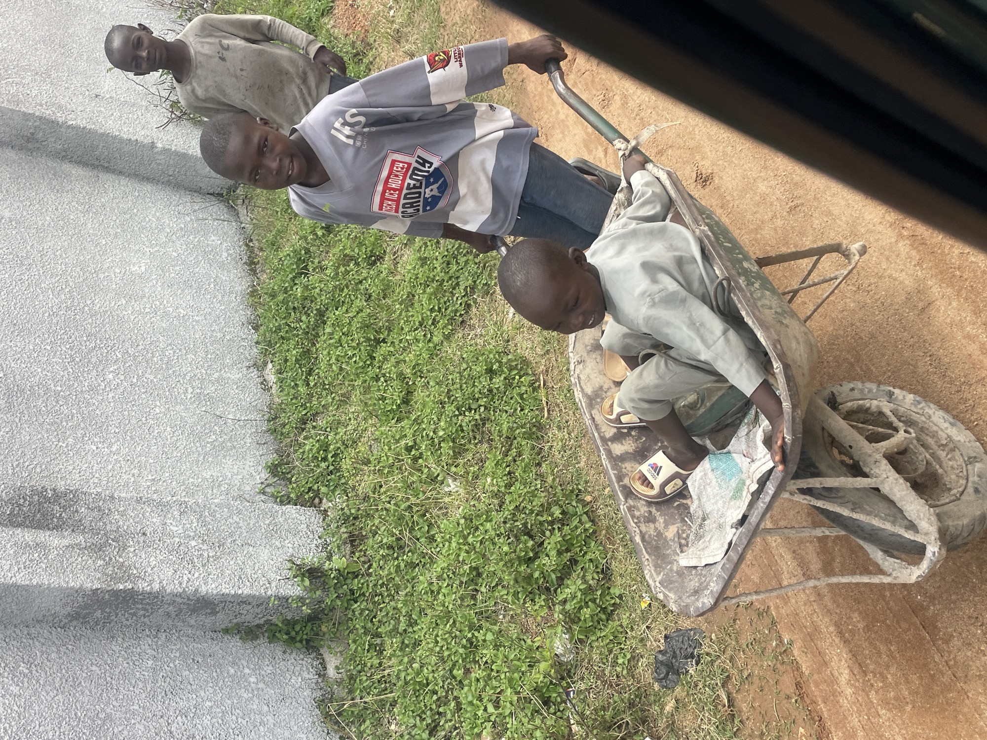 In Jos, Nigeria, two young boys are pictured in an outdoor setting. One boy, wearing a grey long-sleeved shirt with "IFS CZECH ICE HOCKEY ACADEMY" printed on it, is standing and holding the handles of a wheelbarrow. The other boy, seated inside the wheelbarrow, is wearing a light blue long-sleeved shirt. The wheelbarrow appears to be made of metal and is filled with some debris, including newspaper. A third boy, wearing a grey long-sleeved shirt, is visible in the background, standing near a rough-textured, light-colored wall. The ground is covered in dirt and some patches of green vegetation. The image captures a moment of interaction between the children and their environment.