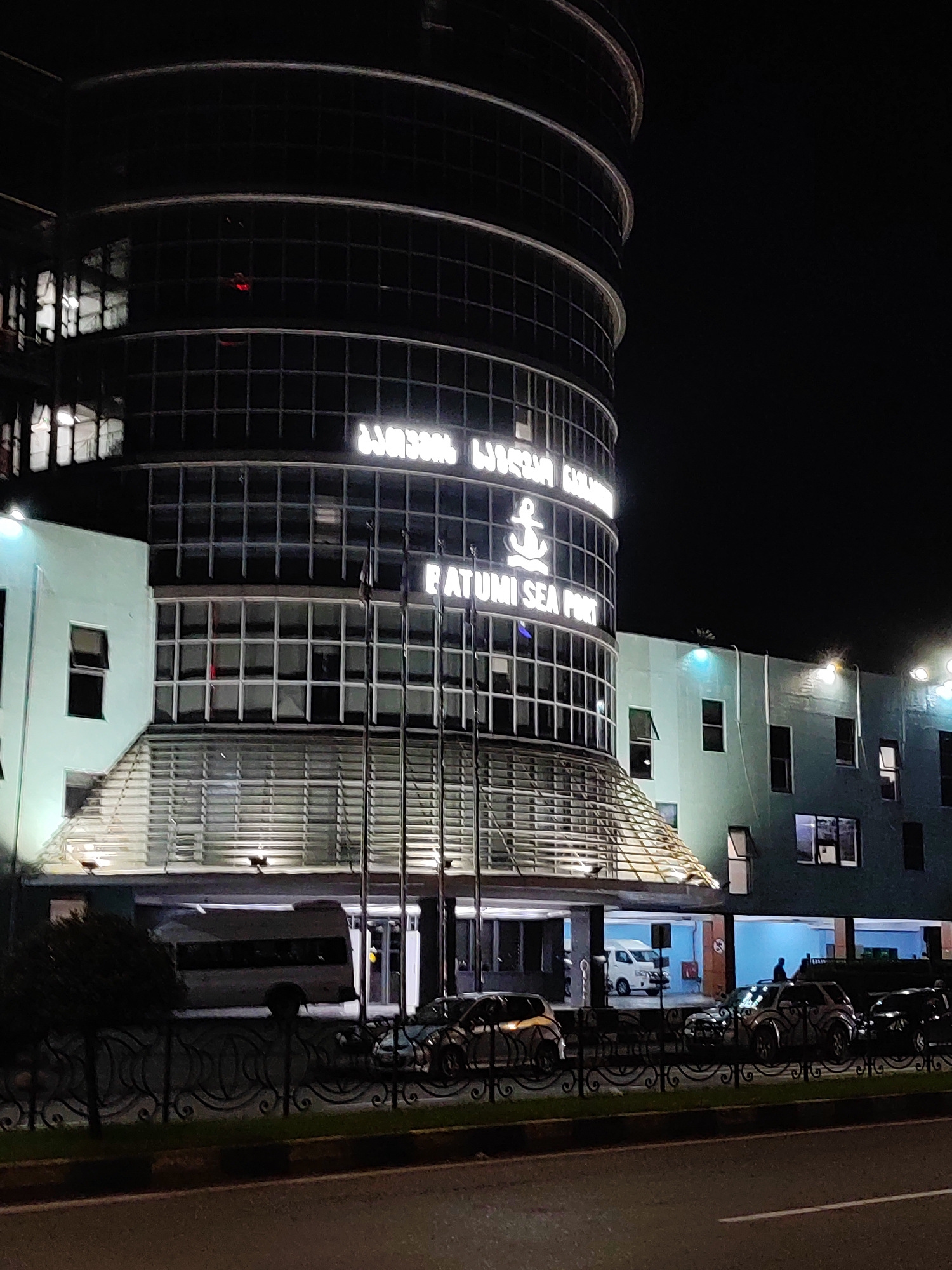 The image captures a nighttime view of a building with a curved, multi-story facade. The building is illuminated, with glowing signage that appears to read "BATUMI SEA PORT" with an anchor symbol. The facade features numerous windows, many of which are lit from within. The lower section of the building has a lighter green color and an angled glass roof structure above an entrance. In front of the building, there is a black metal fence and a road with several parked cars. A white van is visible under the entrance overhang. The sky is dark, indicating it is nighttime. The scene suggests a functional building, possibly a port authority or administrative office, at night.