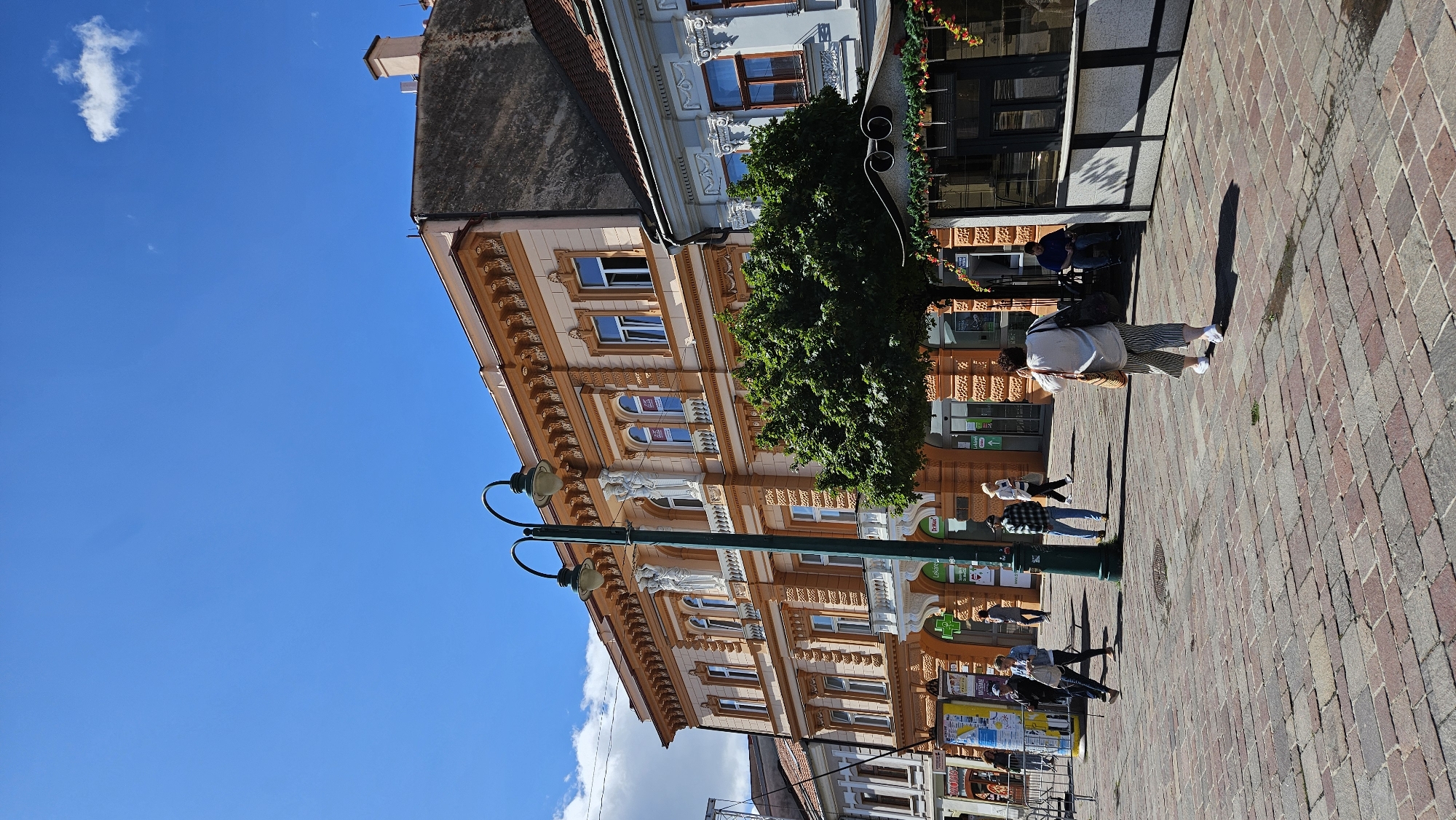 The image shows a wide shot of a city square in Prešov, Slovakia, on a sunny day with a blue sky and scattered clouds. The square is paved with cobblestones and features several people walking. In the foreground, a woman in striped pants and a white top walks away from the camera. To the left, a green lamppost stands prominently in the square.
The background is dominated by historic buildings. The main building on the right is a multi-story structure with ornate orange facades, arched windows, and decorative elements along the roofline. A large green tree partially obscures the lower floors of this building. To the right of this orange building, a lighter colored building with blue accents is visible. Several shops and businesses appear to be located on the ground floor of these buildings, indicated by storefronts and signage. Some people are seen near these establishments. To the far left, a metal fence and more signage are visible. The overall impression is a vibrant urban environment with architectural character and pedestrian activity.