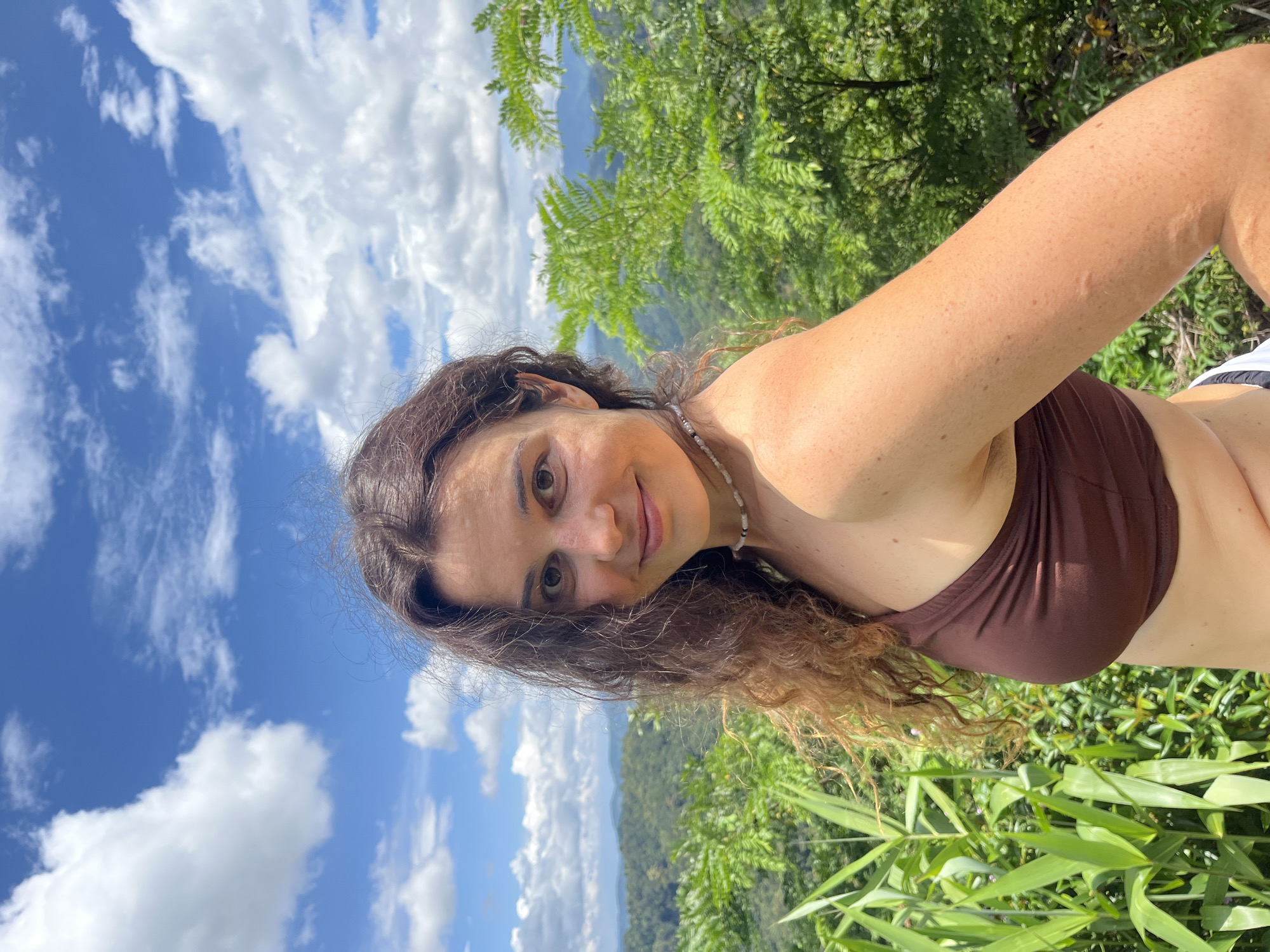 A medium shot captures a woman standing outdoors in Son Dien, Vietnam. She is facing the camera, with her head slightly tilted to her left. The woman has long, curly brown hair and is wearing a brown bikini top. A beaded necklace is visible around her neck. She has a light skin tone and is smiling subtly.
Behind her, a lush green landscape unfolds, with dense foliage and rolling hills covered in trees receding into the distance. The sky above is a vibrant blue, dotted with fluffy white clouds, suggesting a clear, sunny day. The lighting indicates it is daytime. The background elements suggest a natural, possibly mountainous or hilly, environment.