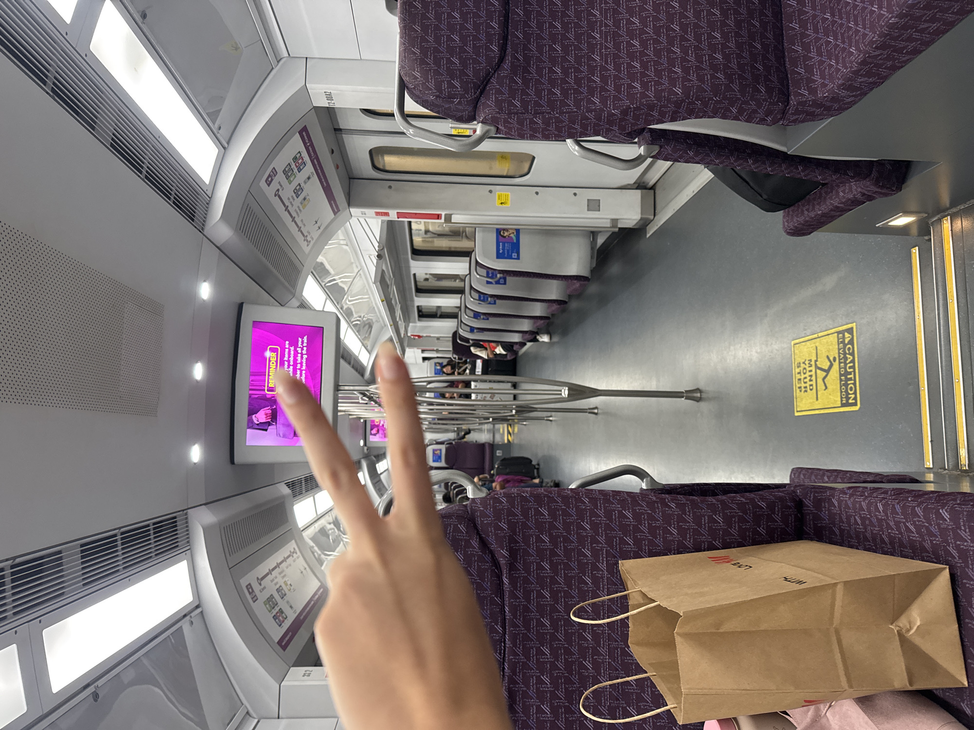 The image shows the interior of a train carriage, likely part of the KLIA Ekspres or KLIA Transit service in Sepang, Malaysia, based on the context of the train's design and its connection to the airport. In the foreground, a person's hand is visible, making a peace sign gesture. To the left, a brown paper shopping bag, with "LOVE WITH H&M" printed on it, rests on a seat upholstered in a dark purple fabric with a geometric pattern. The seats are arranged in a forward-facing configuration along the side of the carriage. The aisle of the train is visible, with more seats and luggage further down. A digital screen mounted on the ceiling displays a pink advertisement. The train carriage is well-lit by overhead fluorescent lights and features metal support poles. A yellow caution sign with an image of a person stepping down is affixed to the floor near the steps. The overall environment appears to be clean and orderly.