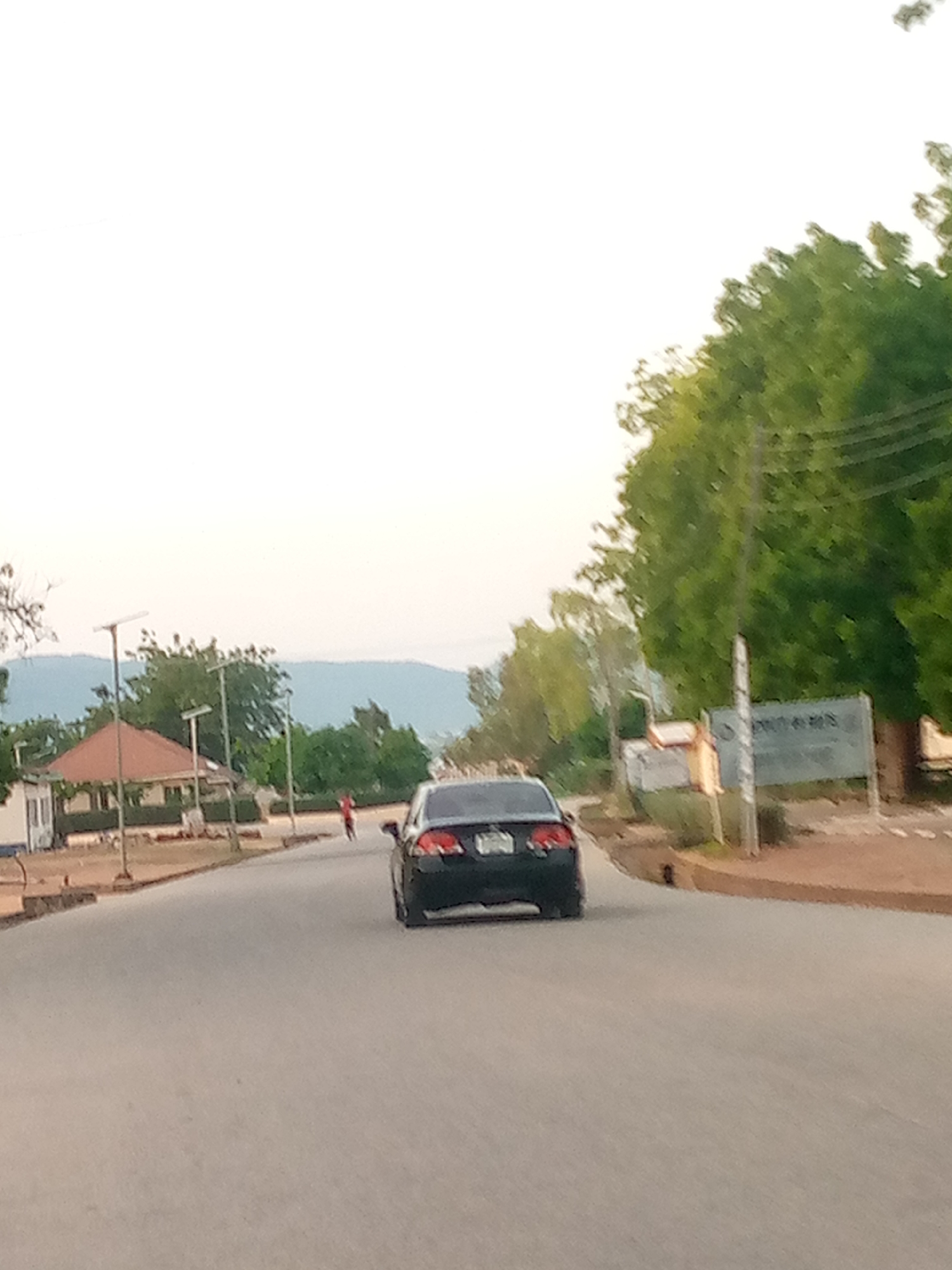 The image depicts a street scene in Jalingo, Nigeria. In the foreground, a dark-colored sedan is driving away from the camera on a paved road. To the left of the road, there is a small building with a red-roofed gazebo-like structure. Several lampposts are visible along the side of the road, some with solar panels. In the middle ground, a lone figure, appearing to be a person wearing red clothing, is standing on the side of the road. Further in the background, dense green foliage and trees are present, with a hazy mountainous landscape visible in the distance. A sign with text, though largely unreadable due to blur, is present on the right side of the image. The sky is overcast and pale.