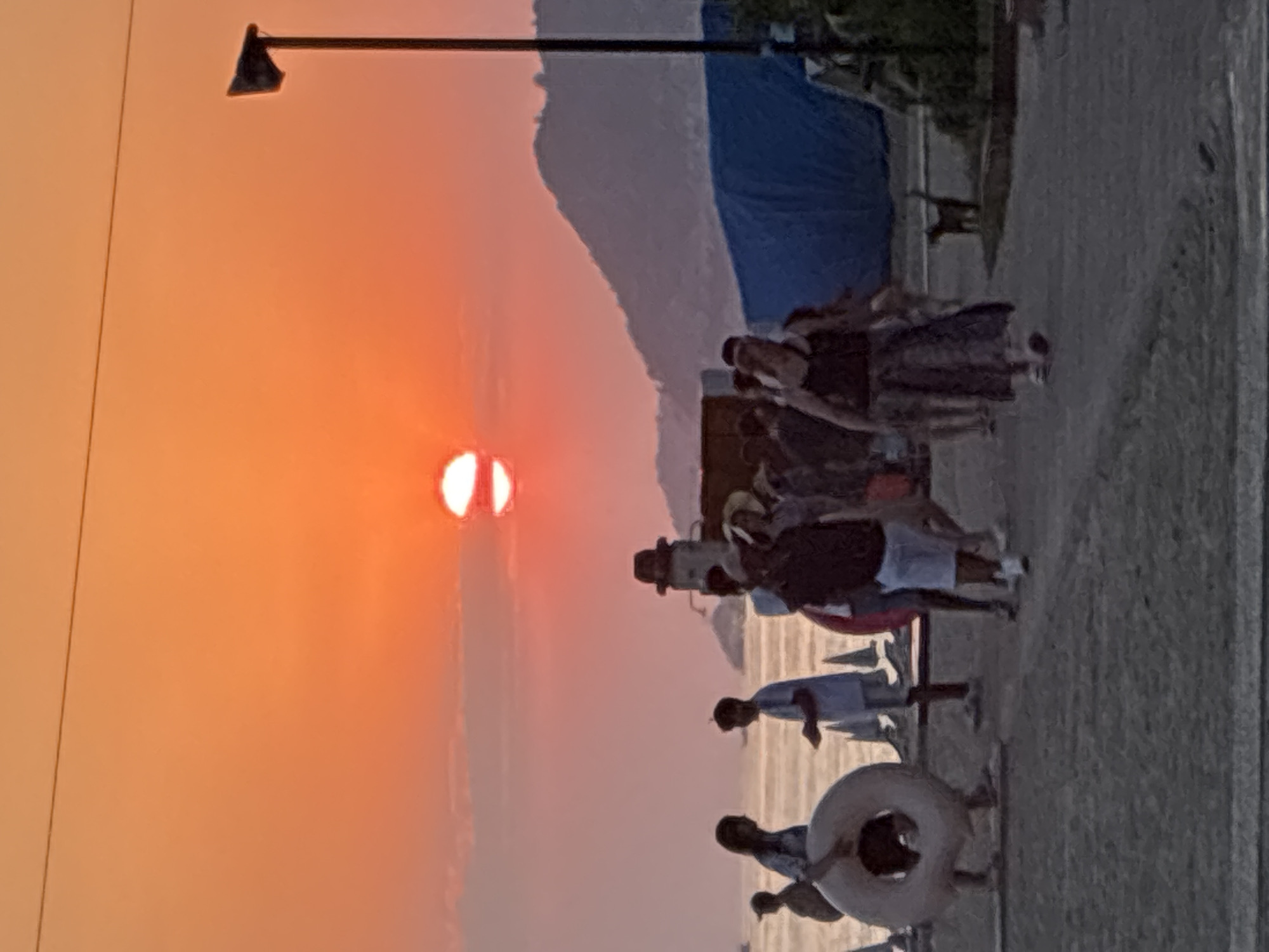 This image depicts a sunset scene near the city of Alanya, Turkey. The sun, a bright orb partially obscured by a thin cloud, hangs low in the sky, casting an orange and pink glow across the horizon. In the foreground, a group of people are walking along a paved area that appears to be a promenade or walkway. Some individuals are holding a large, inflatable ring. In the background, a mountain range is visible, silhouetted against the colorful sky. A tall lamppost stands to the right of the frame. A small, dark-colored dog is present in the lower right corner of the image. The overall atmosphere is that of a relaxed evening by the sea.