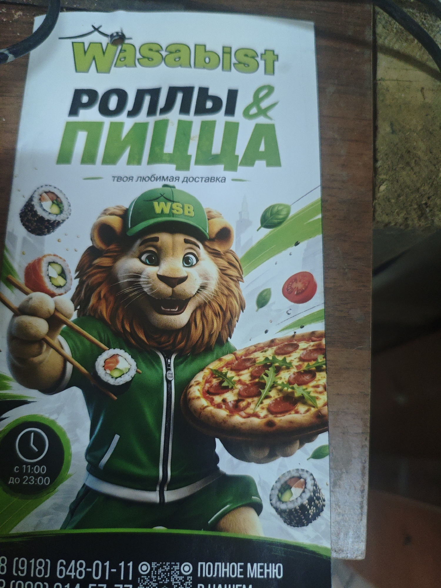 The image shows a flyer for a food delivery service named "Wasabist" that offers rolls and pizza. A cartoon lion character, wearing a green cap with "WSB" on it and a green tracksuit, is depicted holding chopsticks with sushi and a pizza. The flyer also displays the operating hours as "from 11:00 to 23:00" and a phone number starting with "8 (918) 648-01-11". There is also a QR code and the text "ПОЛНОЕ МЕНЮ" (full menu). The flyer has a white background with green and orange accents, and it appears to be placed on a wooden surface. The text indicates that the service is located in Armavir, Russia, as suggested by the "918" area code commonly associated with the Krasnodar Krai region, which includes Armavir.