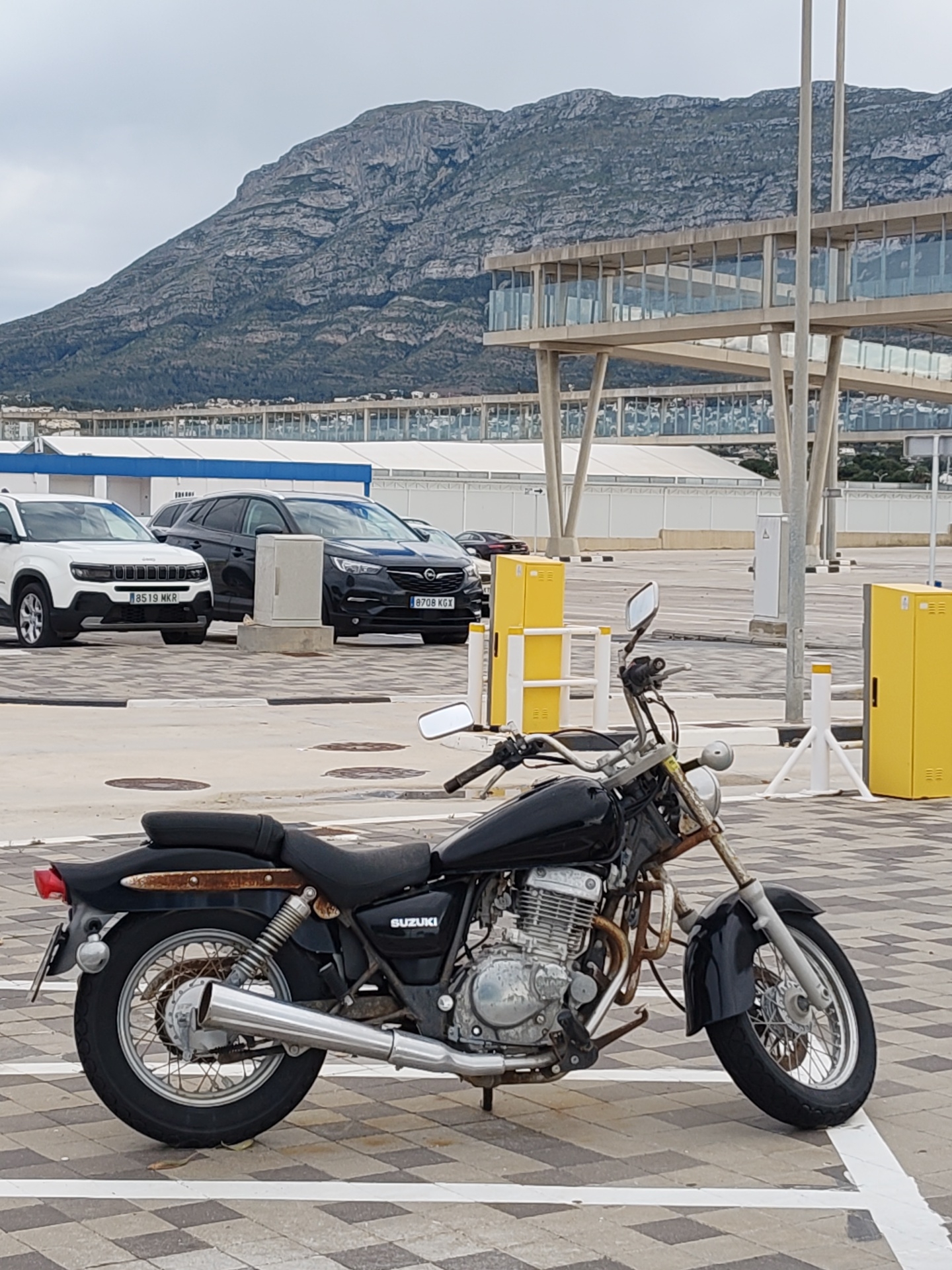 The image is taken in what appears to be a parking area. A black Suzuki motorcycle is prominently positioned in the foreground. In the background, there are several parked cars including a white Jeep with license plate "8519 MKR" and a black Opel with license plate "8708 KGX". Behind the cars, there is a multi-story structure with glass windows supported by steel columns. In the far background, a large mountain is visible under an overcast sky. The ground is paved with square tiles. The presence of cars registered in Spain and a building style consistent with the area suggest the image was taken in the city of Dénia, Spain.