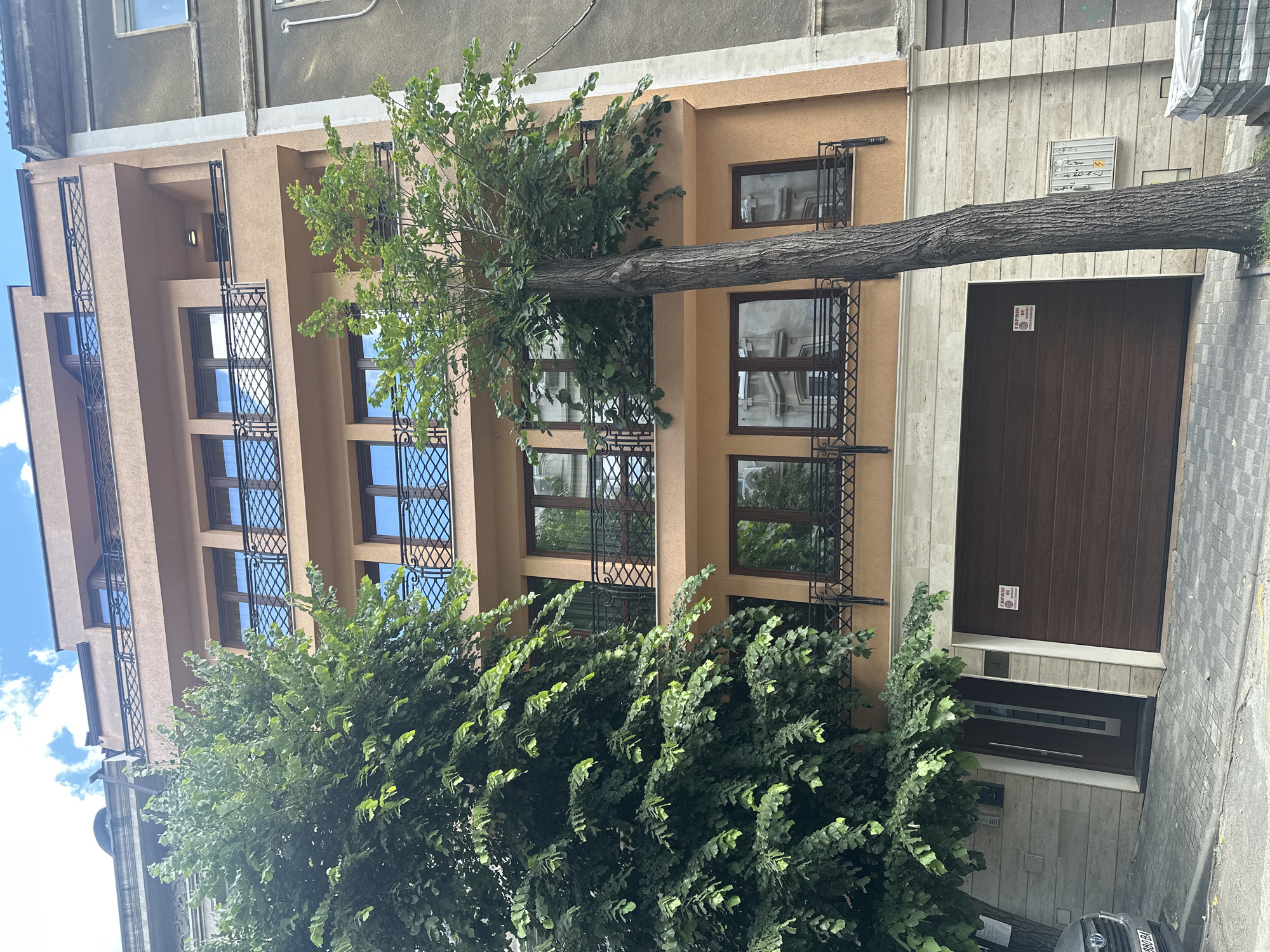 In an exterior, eye-level, full shot of a multi-story building in Unknown, Unknown, a tan, modern-style apartment building stands next to a gray, older building. Two mature trees with lush green foliage are positioned in front of the tan building, with one on the left side of the frame and another in the center, its trunk thick and textured. The tan building features multiple floors with rows of windows, each adorned with black metal railings. The ground floor has a large brown garage door and a dark brown entrance door with a glass panel. The building's facade is a warm tan color, with a beige stone base. The sky above is partly cloudy with white clouds against a blue backdrop. The pavement in front of the building is made of gray interlocking stones, and a portion of a dark-colored car is visible on the left. A sign in Cyrillic, stating "GARAGE NOT WORKING" with a warning symbol, is affixed to the garage door.