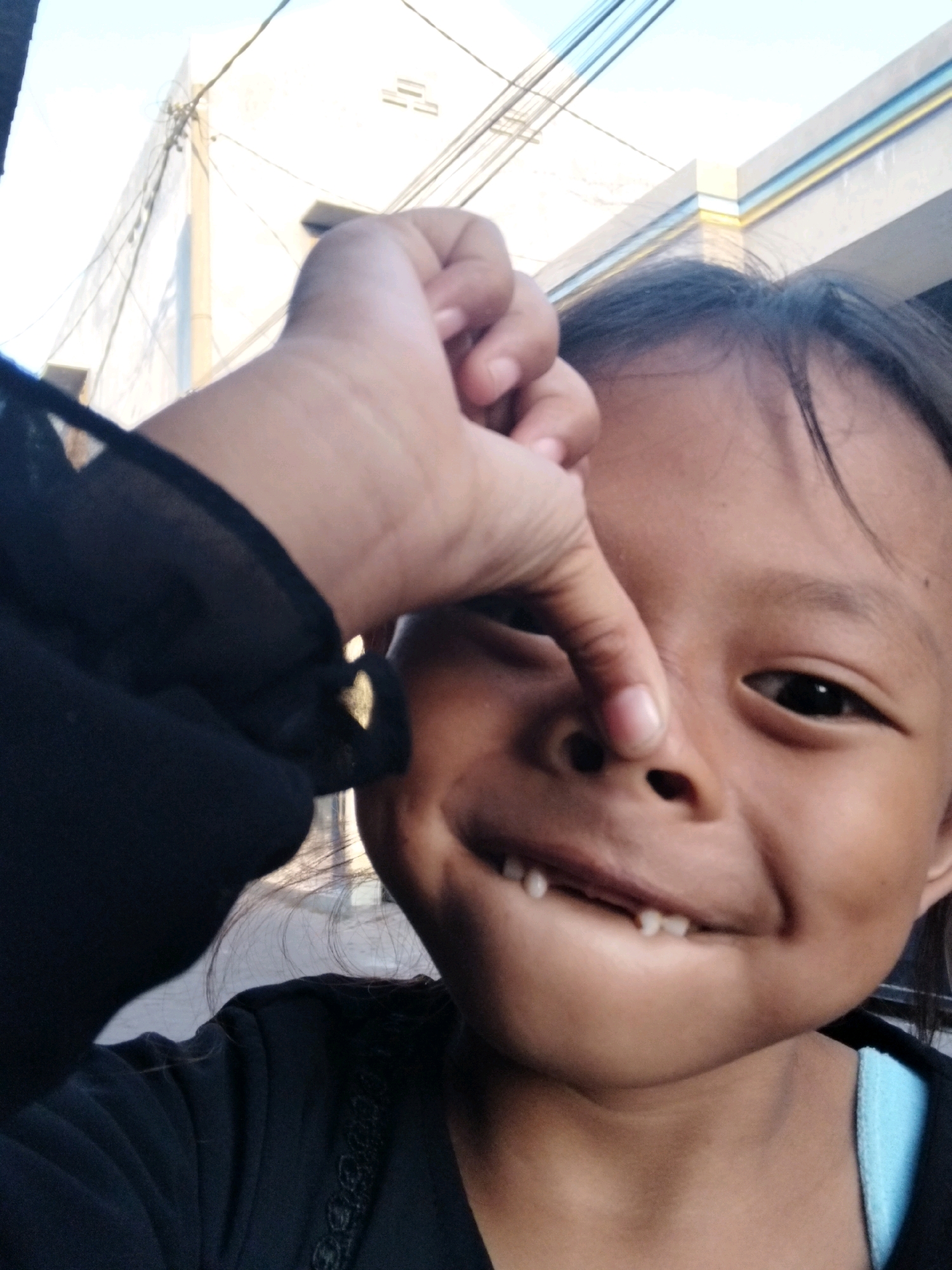 This is a close-up selfie of a young child with dark hair and brown skin, taken outdoors during the day. The child is looking towards the camera with a playful expression, their mouth slightly open, revealing a few visible teeth. A hand, presumably the child's, is reaching up to pinch their nose. The child is wearing a black top with some decorative trim. In the background, a building with white and blue detailing and electrical wires can be seen, suggesting an urban or semi-urban environment. The lighting suggests it's daytime, likely morning or afternoon, with bright sunlight. The overall mood is lighthearted and candid. No text is visible in the image. The location context is Kepuh Kiriman, Indonesia.