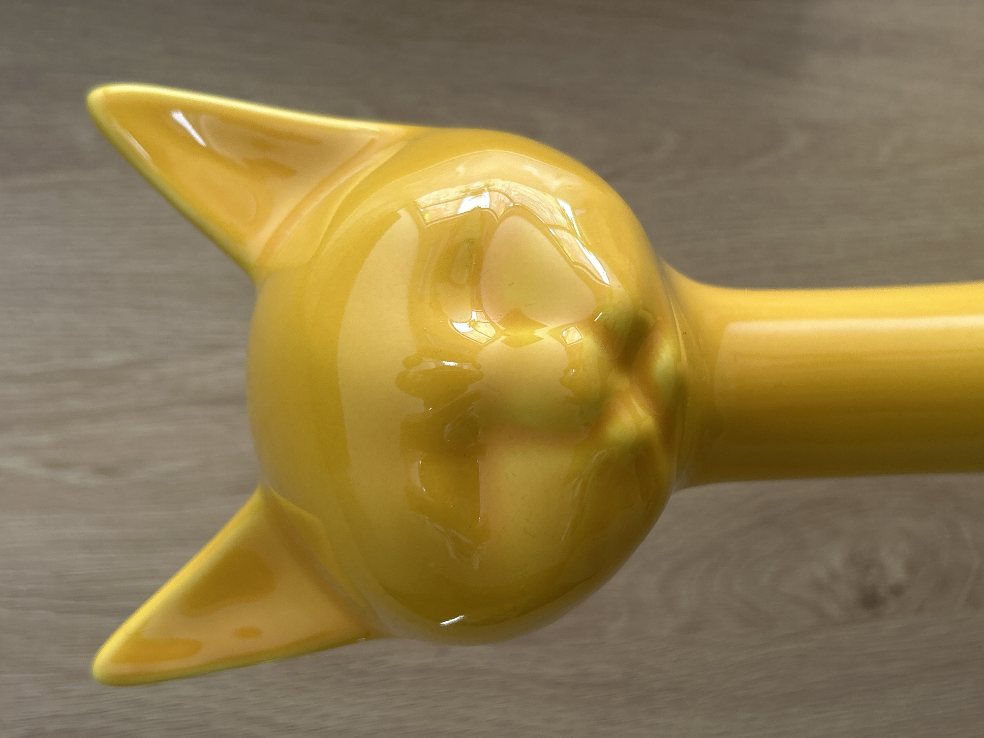 A close-up shot shows a yellow, glazed ceramic cat figurine's head and part of its body lying on a light brown, wood-patterned surface. The cat has pointed ears and a round head with a subtle, somewhat obscured facial feature. The glaze creates shiny highlights. The background is out of focus, showing more of the wood-patterned surface. The location in Sofia, Bulgaria, is not evident in the image.
