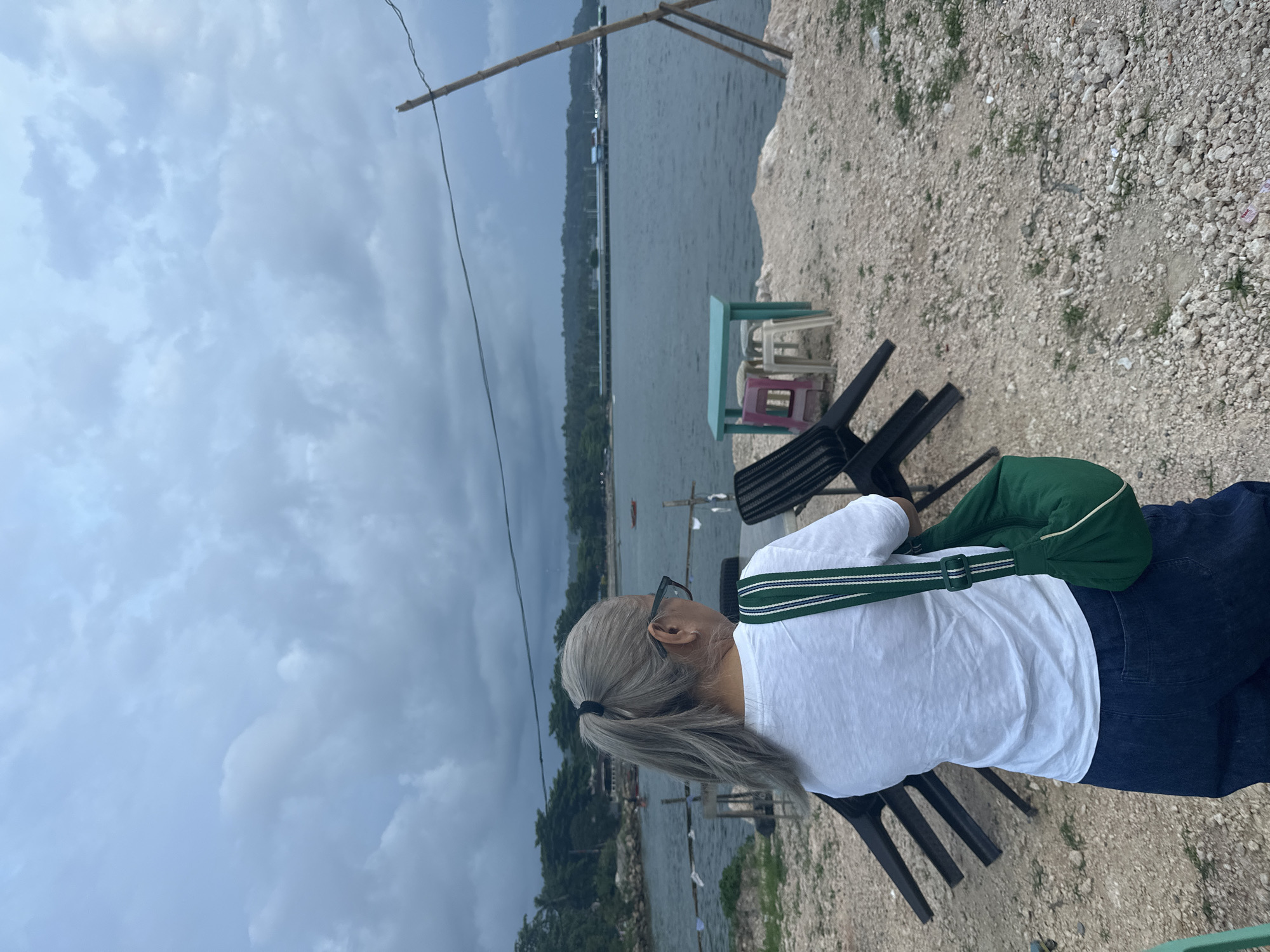 A woman with grey hair pulled into a ponytail, wearing glasses, a white t-shirt, and dark blue jeans, stands facing away from the camera, looking towards a body of water. She is carrying a green bag with white and green straps over her right shoulder. In the foreground, several black chairs are stacked or overturned on a rocky, sandy ground. In the middle ground, a small table with colorful plastic stools sits near the water. The background features the sea under a cloudy sky, with a distant shoreline lined with trees and structures, possibly a pier or buildings. A makeshift bamboo pole with wire attached is visible in the right portion of the image, suggesting a rural or undeveloped area. The overall scene appears to be a coastal location in Samal, Philippines, likely a casual outdoor seating area overlooking the sea.