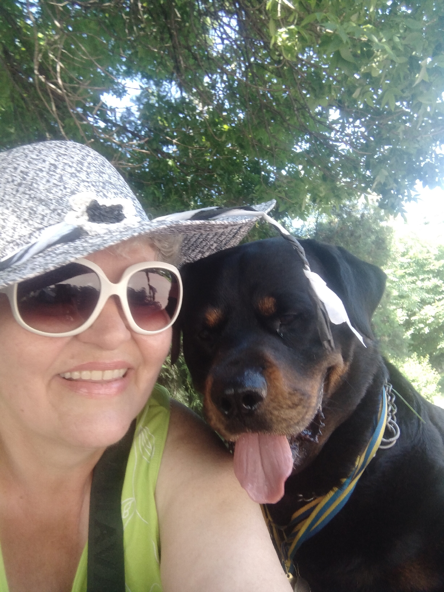 A woman wearing a white and grey patterned hat and large white sunglasses smiles at the camera. She has fair skin and is wearing a light green top. Next to her, a Rottweiler dog, wearing a blue and yellow striped collar, rests its head. The dog's tongue is hanging out. They are outdoors, with green trees and foliage visible in the background, suggesting a park or natural area. The lighting indicates it is daytime, likely sunny. There is no information to determine the specific location in Horishni Plavni, Ukraine.