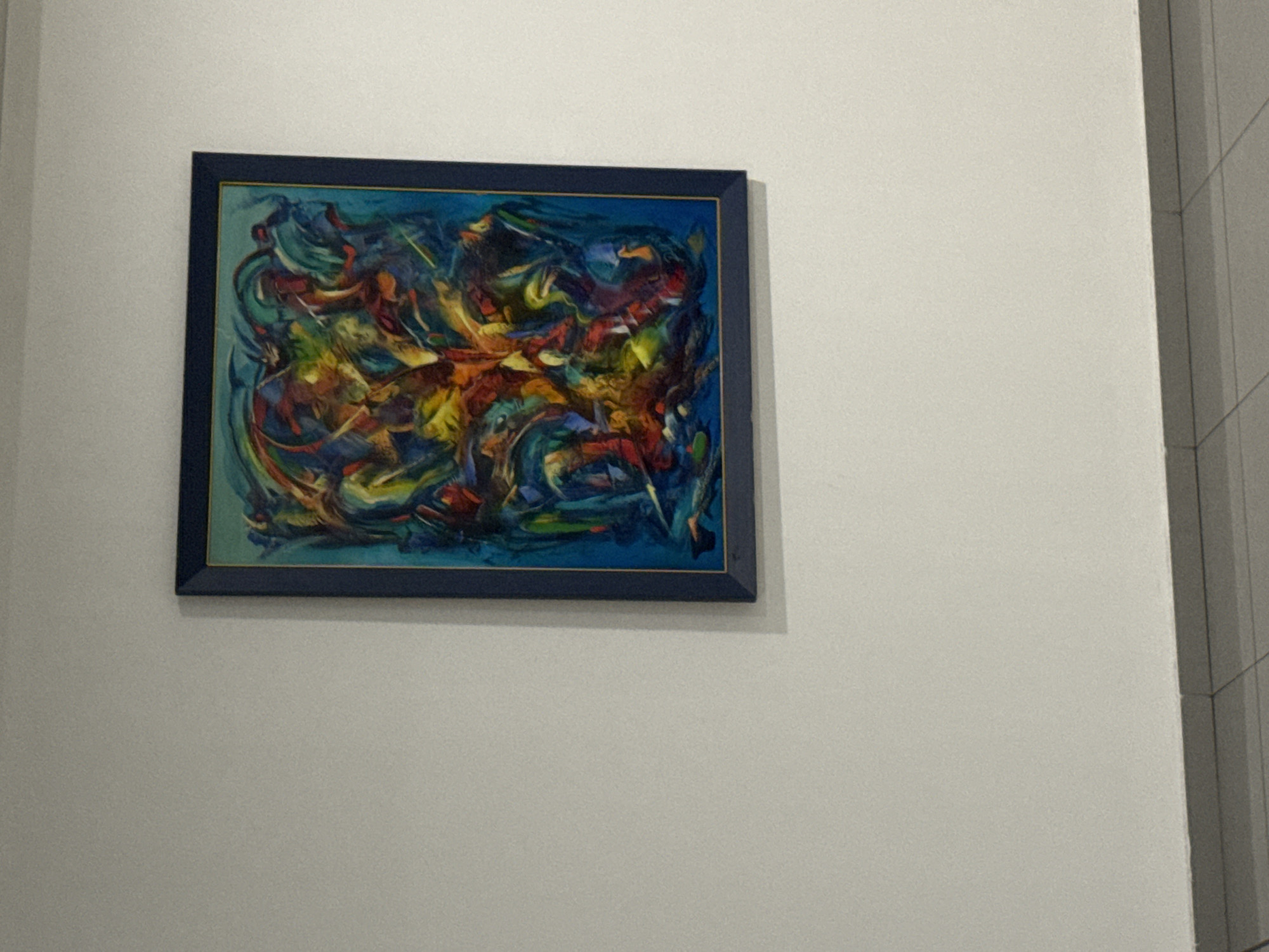 A framed abstract painting hangs on a white wall. The painting has a blue frame. The artwork features a combination of blue, red, yellow, and green hues, depicting swirling shapes and lines. The environment appears to be an indoor space, potentially a gallery or a room within a building, but without additional context, the specific location in Sofia, Bulgaria cannot be determined.