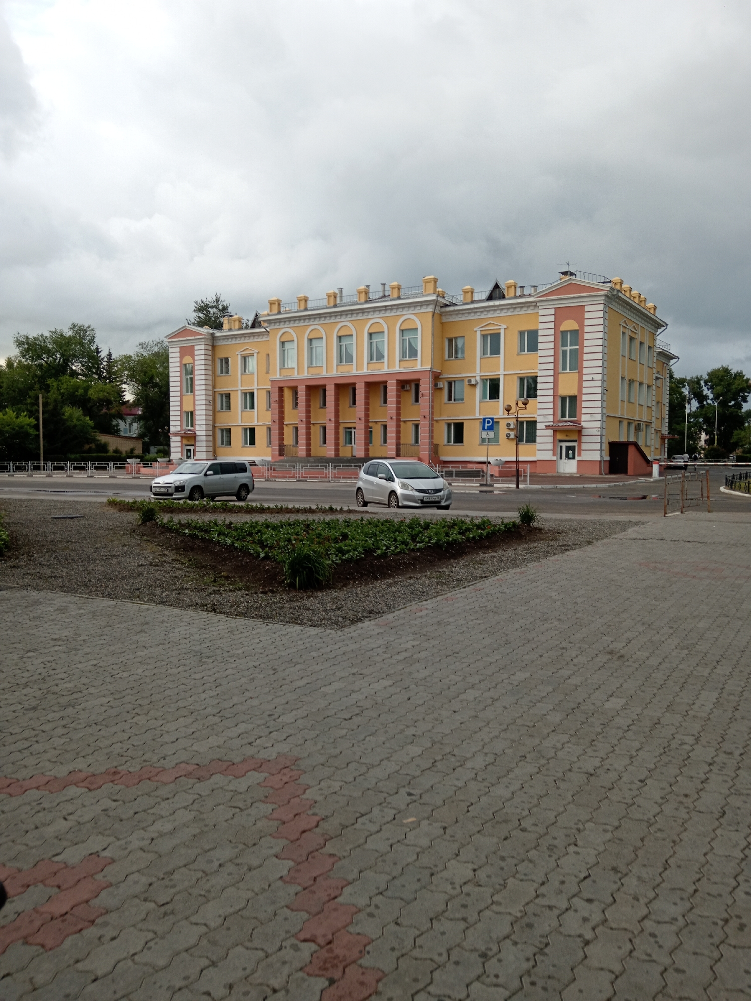 The image depicts a street scene in Belogorsk, Russia, under a cloudy sky. In the foreground, a paved walkway with a distinct red and gray interlocking brick pattern is visible. Beyond this, a circular flower bed with green plants and mulch separates the walkway from a paved road. Two cars are present on the road: a silver compact SUV in the middle distance and a white car partially visible on the right edge of the frame.
Dominating the background is a three-story building with a light yellow facade, accented with pink and white trim. The building features numerous windows and a classical architectural style with decorative elements along the roofline and entrance. A blue and white parking sign is mounted on a pole in front of the building. Trees and foliage are visible to the left and right of the building, suggesting a semi-urban or suburban setting. The overall impression is one of a public space with functional infrastructure and a well-maintained building.