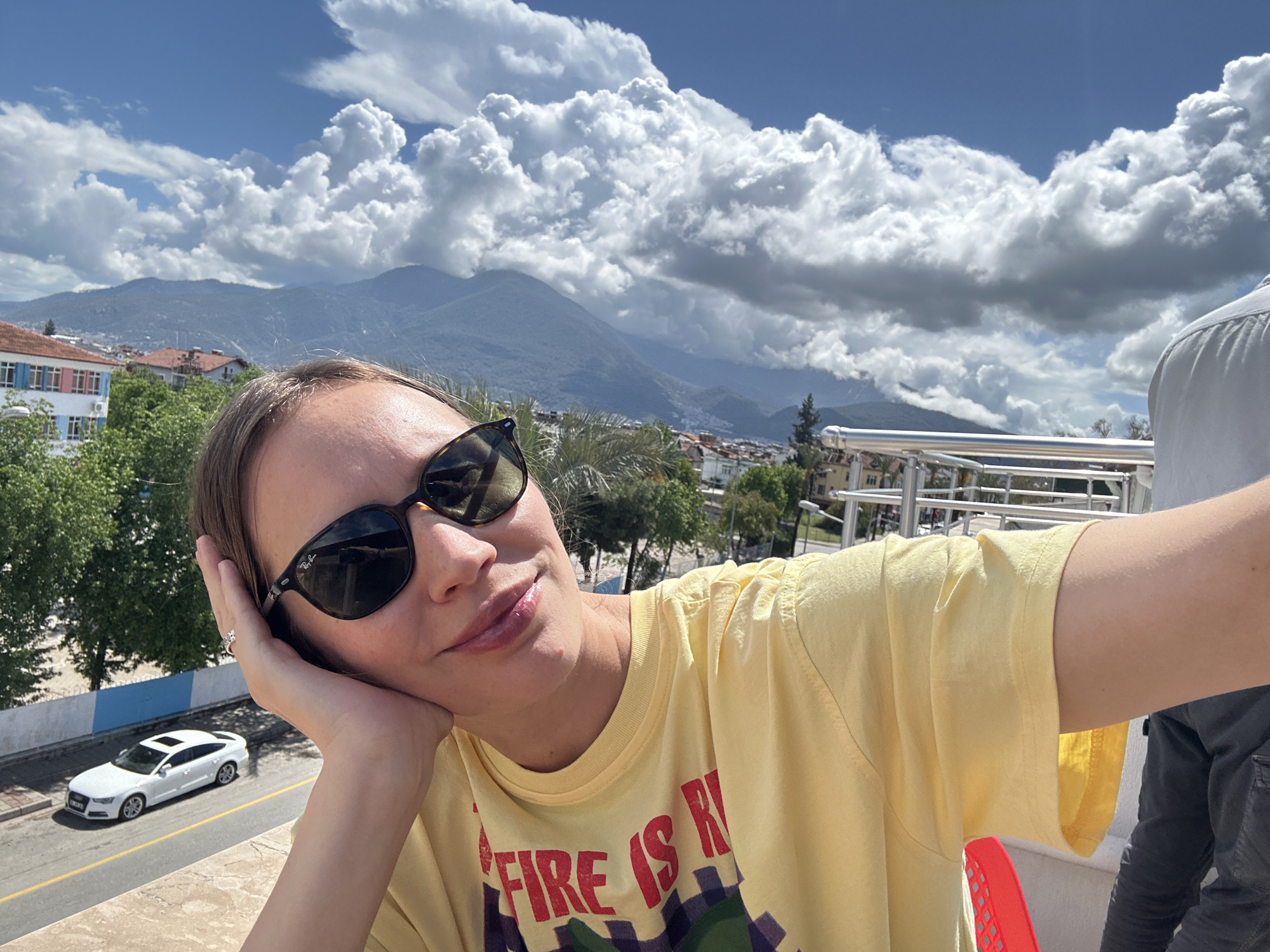 A woman with light skin and short hair poses for a selfie, wearing sunglasses and a yellow t-shirt with a colorful design. She is resting her head on her hand, and a ring is visible on her finger. Behind her, there's a street with a white car parked on it, trees, and buildings. In the distance, mountains rise against a blue sky filled with large, puffy clouds. The scene appears to be in an urban area, specifically Fethiye, Turkey, as suggested by the architecture and mountainous backdrop. Other people can be seen close in the background.