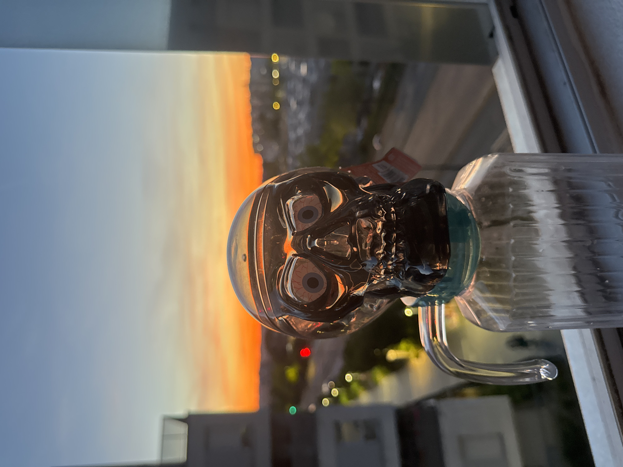 A glass skull object, resembling a skull-shaped pipe, is positioned in the foreground, centered in the frame. The skull has visible, detailed eyes with a red and blue iris. It is attached to a clear, ribbed glass bottle with a curved glass handle. The skull and bottle are illuminated by the warm light of a sunset. In the background, a city skyline is visible under a gradient sky of blue and orange. The city lights create bokeh effects in the distance, suggesting it is dusk. The objects are placed on a windowsill, with a window frame visible to the right of the image.