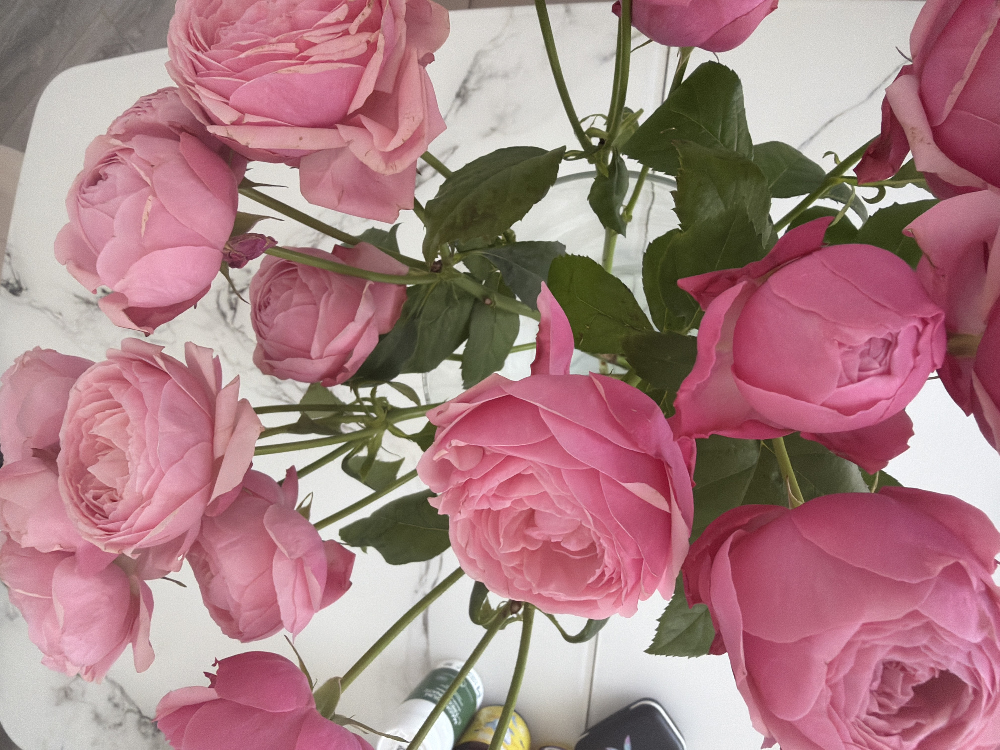 The image shows a close-up, top-down view of a bouquet of vibrant pink roses. The roses are full-bloomed and clustered together, with some buds visible. Their petals are layered and soft, with subtle variations in shade from pale pink to a deeper rose hue. Green leaves and stems are interspersed among the flowers, adding a contrasting natural element. The bouquet is placed on a white surface with a subtle marbled pattern, suggesting it might be a table or countertop. In the background, partially visible, are a few small cylindrical containers, one white with a green label and another with a yellow label, and a dark rectangular object that appears to be a phone or small electronic device. The lighting is bright, illuminating the delicate details of the rose petals. The scene does not contain any identifiable landmarks or features specific to Malinovka, Russia.