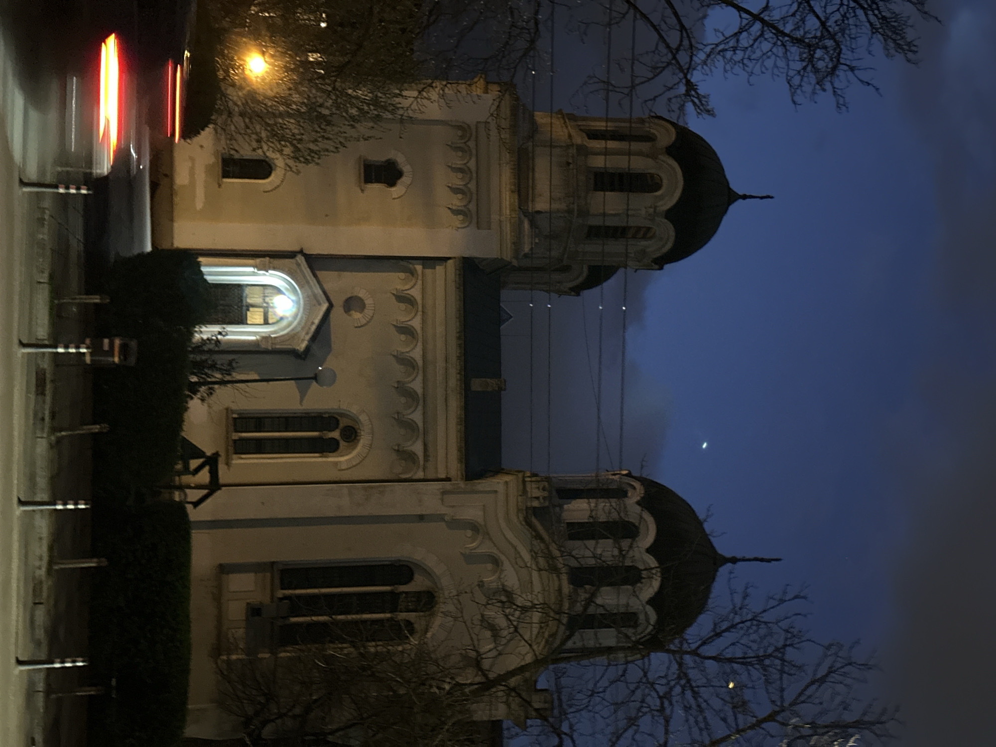 A nighttime street scene in Sofia, Bulgaria shows the Saint George Rotunda Church. The light-colored stone building has two dome-topped towers, and bare tree branches frame the scene. A street runs along the building's left side, with blurry car lights. The sky is a mix of dark blues and grays.