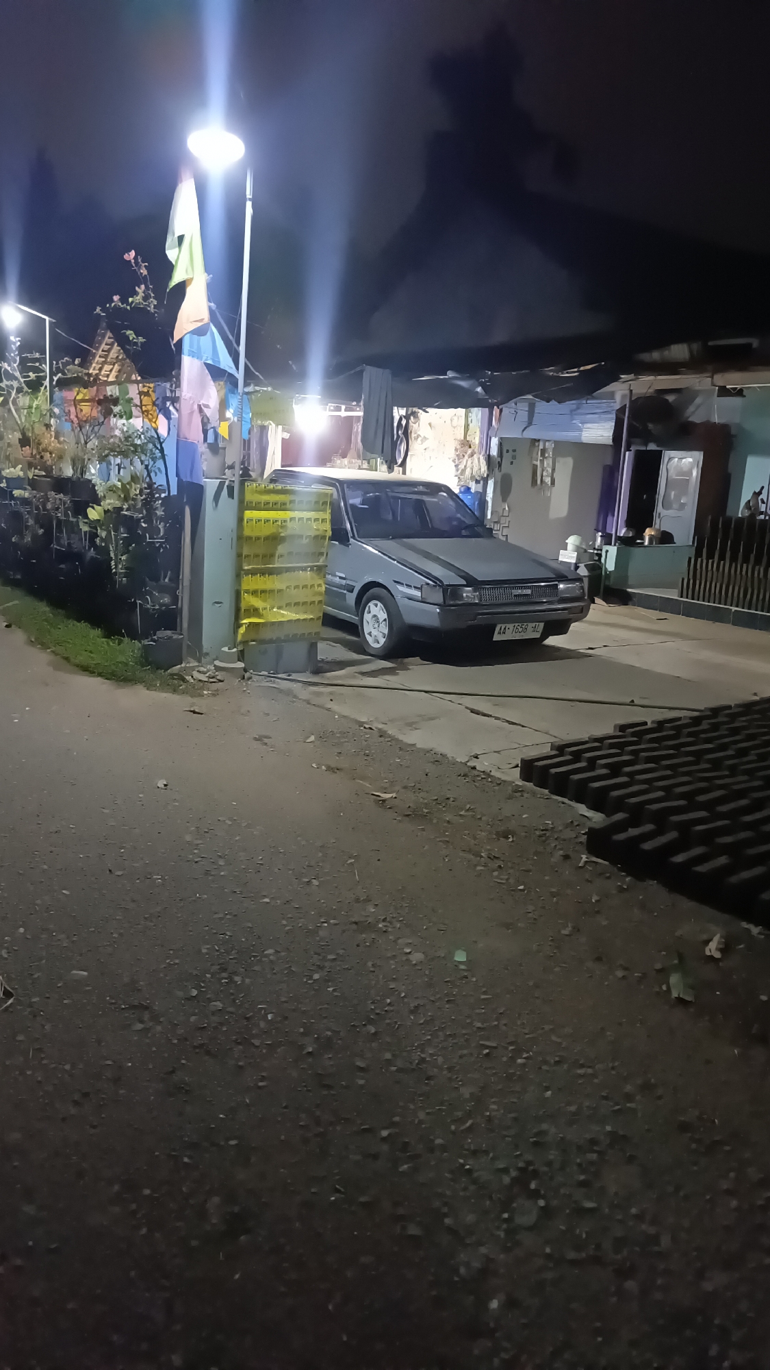 An older model grey sedan is parked on a paved driveway in front of a building in Kebumen, Indonesia. The car has a license plate that reads "AA-1658-HL". To the left of the car is a light pole with several flags and laundry hanging on a clothesline. A stack of yellow crates is positioned next to the light pole. The scene is dimly lit, with a street lamp casting a bright light on the flags and the car. In the foreground, the ground is covered with gravel and some debris. The background shows parts of buildings, with awnings and shadows indicating it is nighttime.
