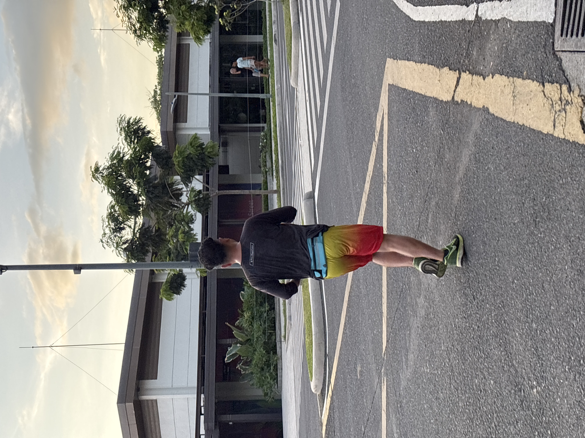 A male runner is seen from behind, running on an asphalt road with yellow and white markings. He is wearing a dark long-sleeved shirt, yellow and red gradient shorts, a blue fanny pack, and green running shoes. In the background, a modern building with a dark brown roof and white facade is visible. Trees and a light pole are also present. The sky is partly cloudy with a warm, sunset glow. The scene appears to be in a park or an open area with pedestrian crossings and landscaped greenery, potentially part of the University of the Philippines Mindanao campus in Davao City, Philippines, given the architectural style and the presence of an antenna array on the building which is characteristic of educational institutions. Two other individuals are visible walking in the distance.