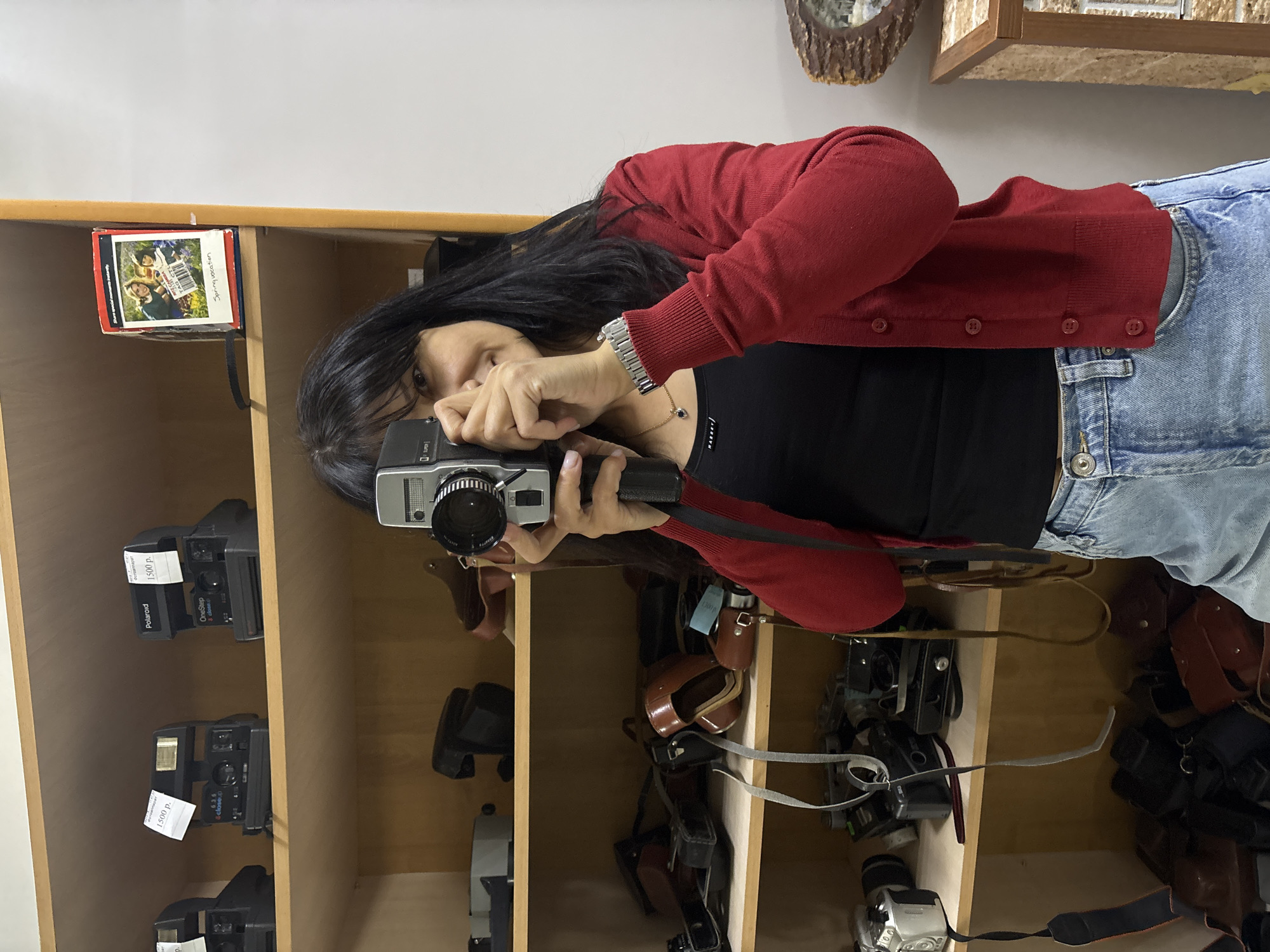 A woman with long dark hair, wearing a red cardigan over a black top and blue jeans, is captured in an indoor setting, possibly a vintage camera store or display. She is actively taking a photograph, holding an older model silver and black camera up to her eye, her left hand grasping the camera and showing a silver wrist watch.
The background features wooden shelves filled with various cameras and accessories. On the left shelves, multiple Polaroid cameras are visible. One is identified as "Polaroid OneStep" and another as "close.p", both bearing price tags of "1500 р." (Russian Rubles). A small box on the top shelf displays the text "Spring vocation" and "Share special moments instantly," along with images of people. The shelves to the right contain more vintage cameras, some nestled in leather cases with straps. The visible pricing in Russian Rubles provides a strong location cue, aligning with the context of Chaikovsky, Russia. The image suggests an appreciation for classic photography equipment and hints at an engaging activity within the store.