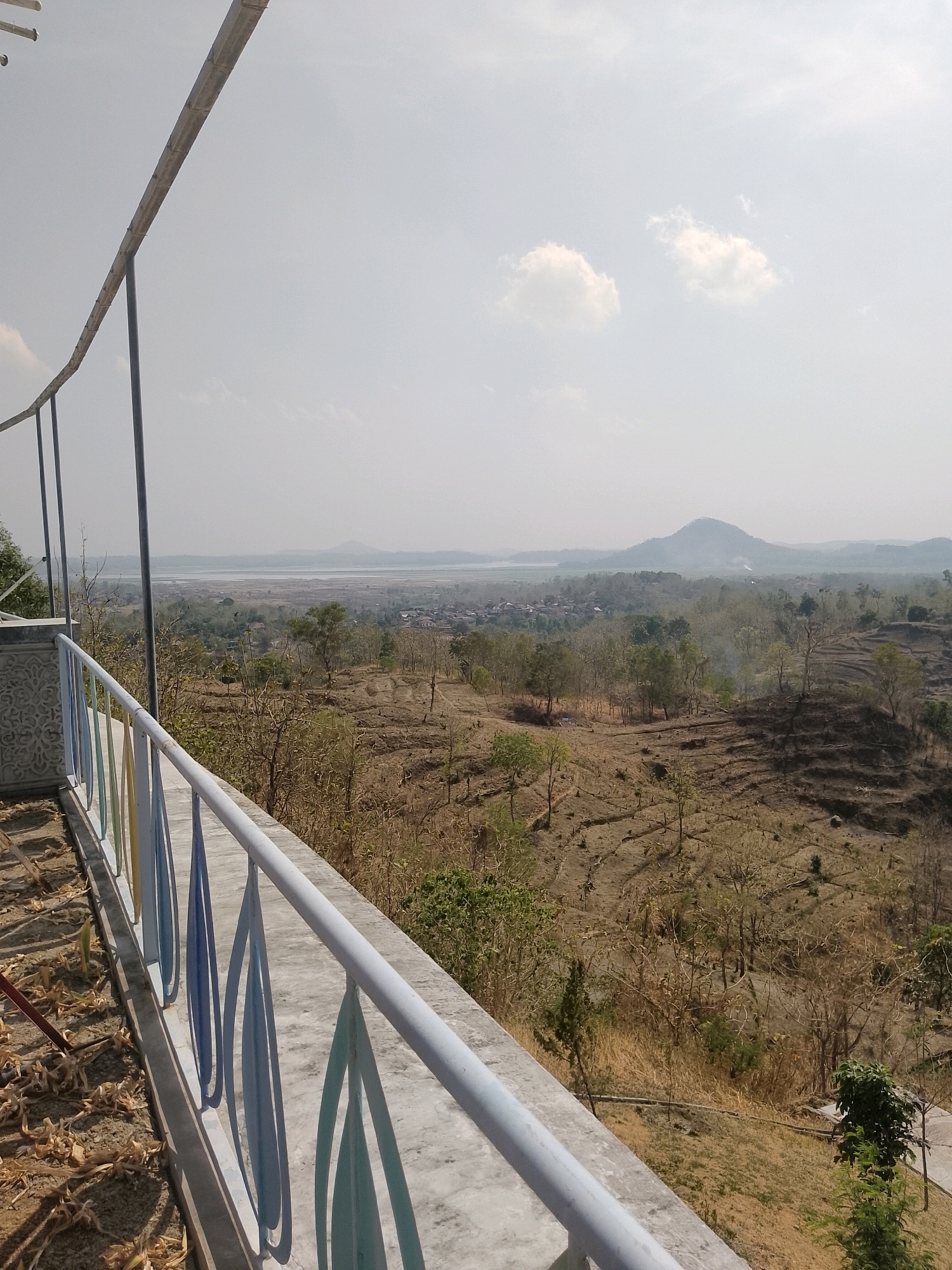 The image shows a panoramic view of a rural landscape, likely in or around Penanggapan, Indonesia, from a high vantage point. In the foreground, a concrete balcony or walkway is visible with a light-colored railing that has decorative, tear-drop shaped elements painted in shades of blue and green. The railing runs diagonally across the bottom left of the frame. The ground on the balcony appears to be concrete, with some dried leaves and stalks scattered on it.
The landscape beyond the balcony is characterized by dry, terraced hillsides dotted with sparse vegetation, consisting primarily of dry grasses and leafless trees, suggesting a dry season. In the mid-ground, a village or settlement is nestled amongst the trees, with buildings visible. Further in the distance, a body of water, possibly a lake or river, stretches across the horizon, bordered by more hills and mountains under a hazy sky. The sky itself is bright and mostly clear, with a few scattered white clouds. The overall impression is one of a dry, arid rural environment with a distant water feature and mountainous terrain.