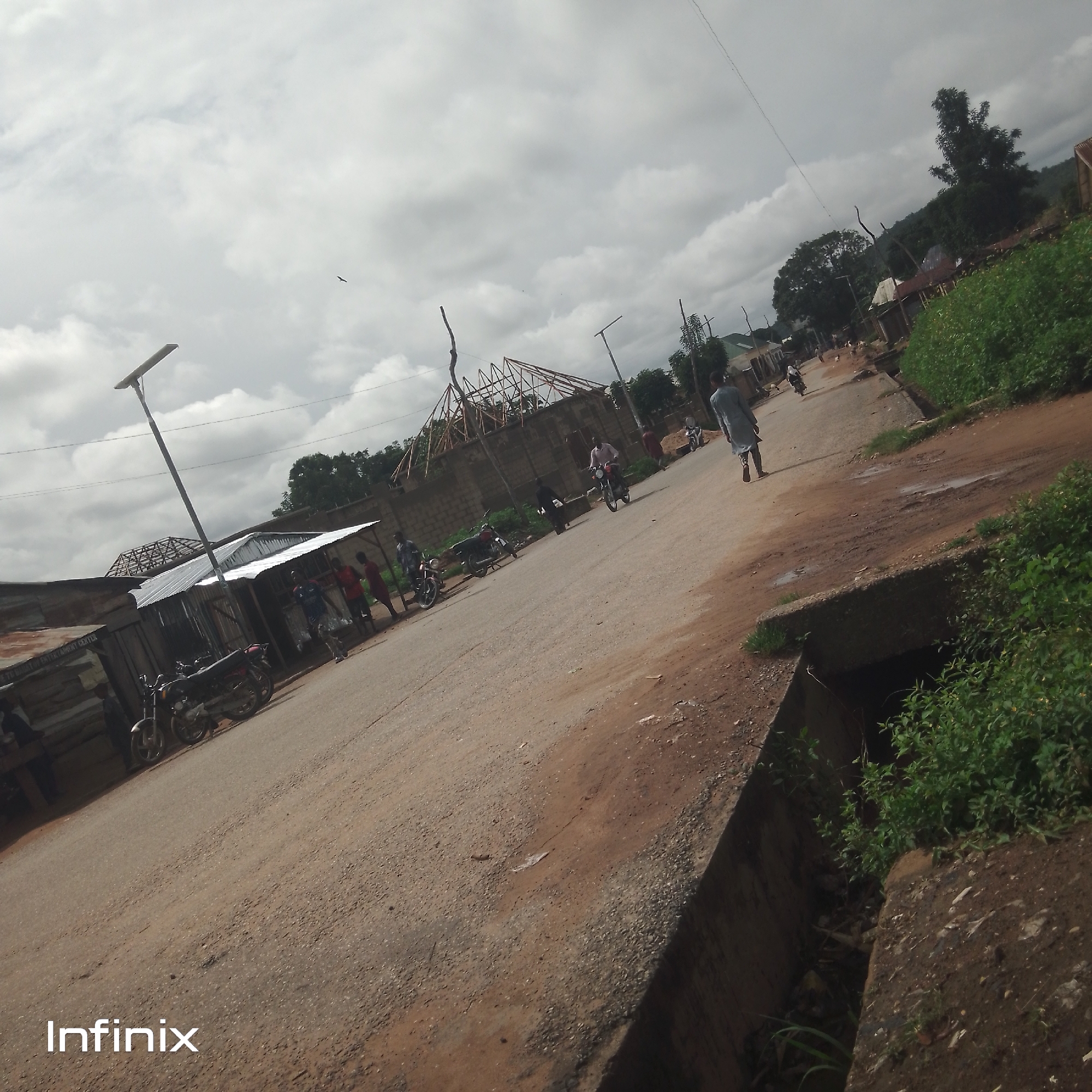 The image depicts a street scene in an outdoor, urban environment. The street is unpaved and appears to be made of dirt and gravel. On the left side of the street, there are several buildings with corrugated metal roofs. In front of these buildings, several motorcycles are parked. A street light with a solar panel is also visible.
People are present in the scene. One person is walking away from the camera down the street. Several other individuals are seen standing near the buildings. A person is riding a motorcycle down the street.
The sky is overcast with clouds. There are trees and vegetation on the right side of the street, and some hills can be seen in the background. A concrete drainage ditch runs along the right edge of the street.
The word "Infinix" is visible in the lower-left corner of the image, suggesting it may be a watermark or a branding element.