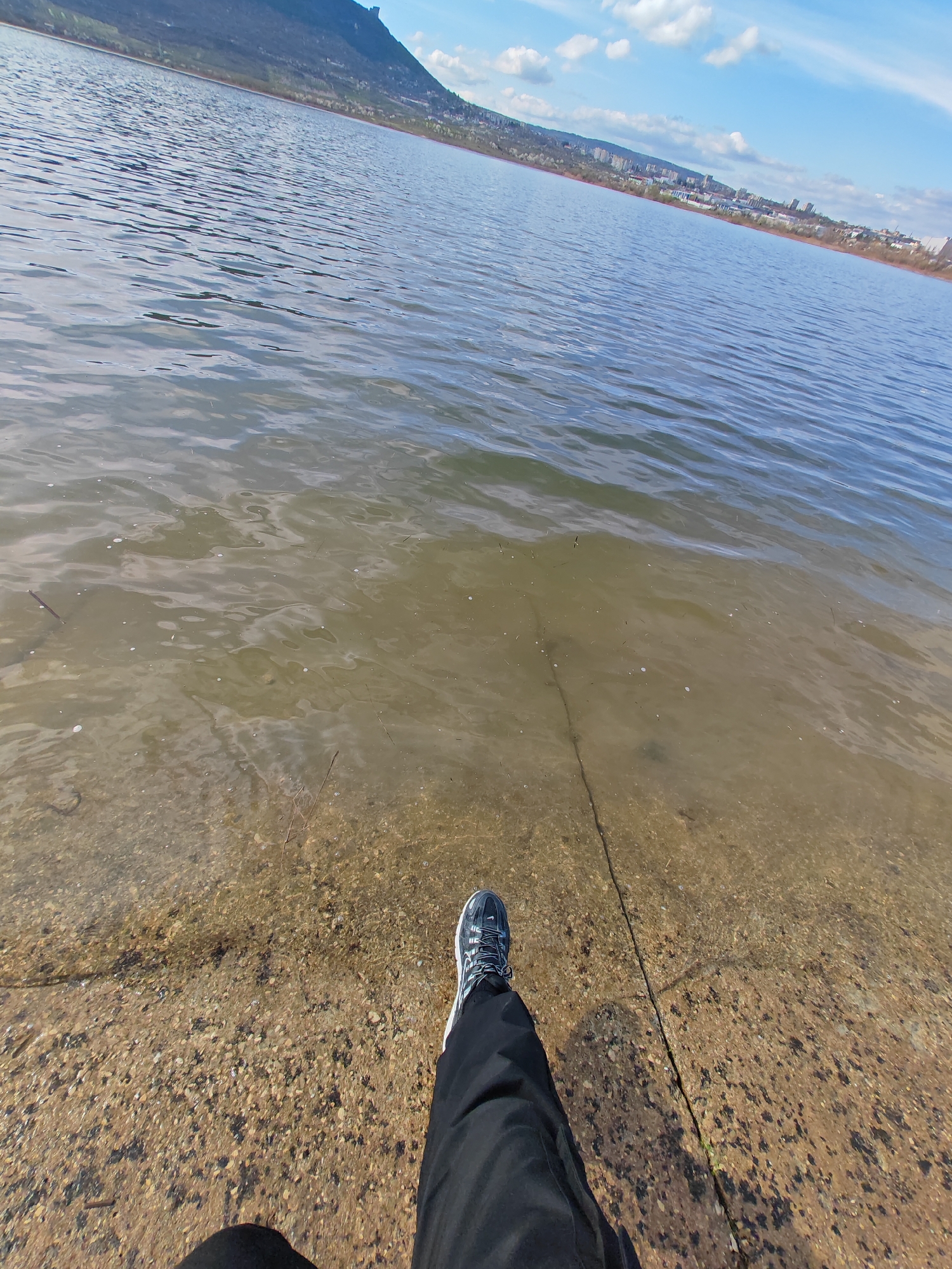 A ground-level, point-of-view shot shows a person standing on a concrete bank at the edge of a body of water. The water is clear near the shore, revealing a gravelly bottom. Ripples are visible on the surface. In the background, a mountain and the skyline of Shumen, Bulgaria can be seen under a blue sky with scattered clouds. The person is wearing a blue and grey athletic shoe and dark pants. A narrow crack is present in the concrete surface where the person is standing.