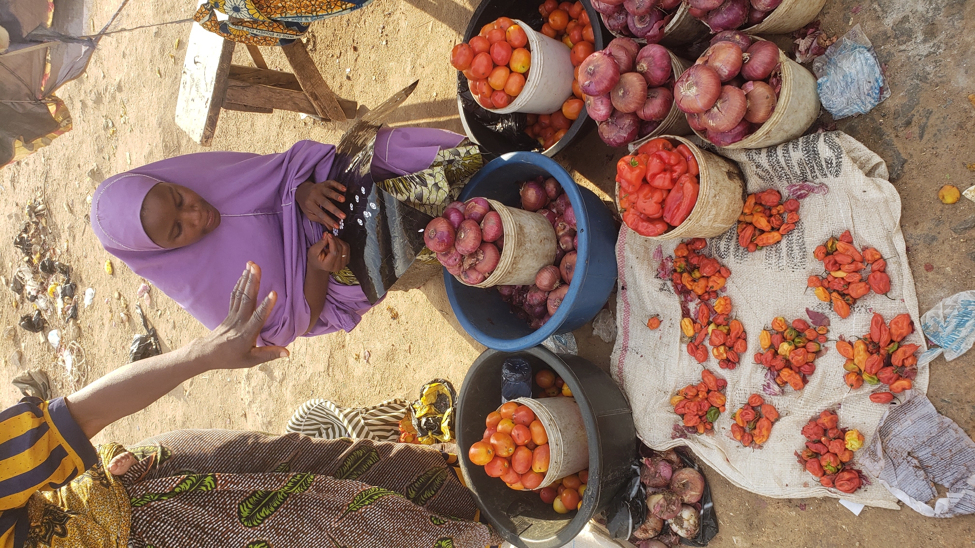 The image depicts a market scene in Kawo, Nigeria. A woman wearing a lilac hijab sits beside buckets and displays of produce. The produce consists of tomatoes, onions, and habanero peppers. Some peppers are laid out on a cloth. A man's hand reaches into the frame from the left. The ground is sandy.