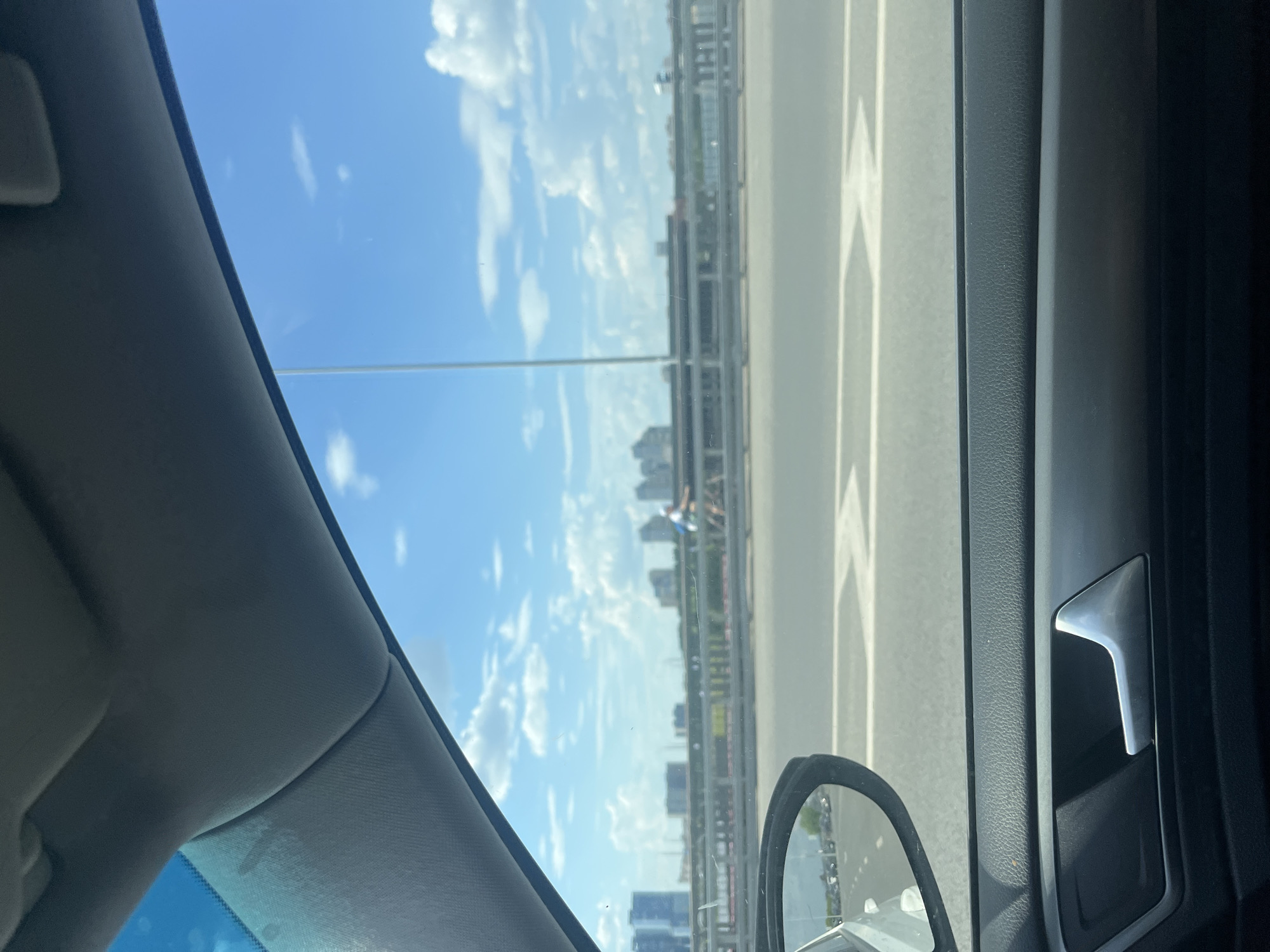 A view from inside a vehicle shows a bright, cloudy blue sky above a cityscape with tall buildings in the distance. A bridge spans across the scene, with a person riding a bicycle on it. The vehicle's interior, including a door handle and part of the dashboard, is visible in the foreground. The reflection in the side mirror shows other vehicles on the road.