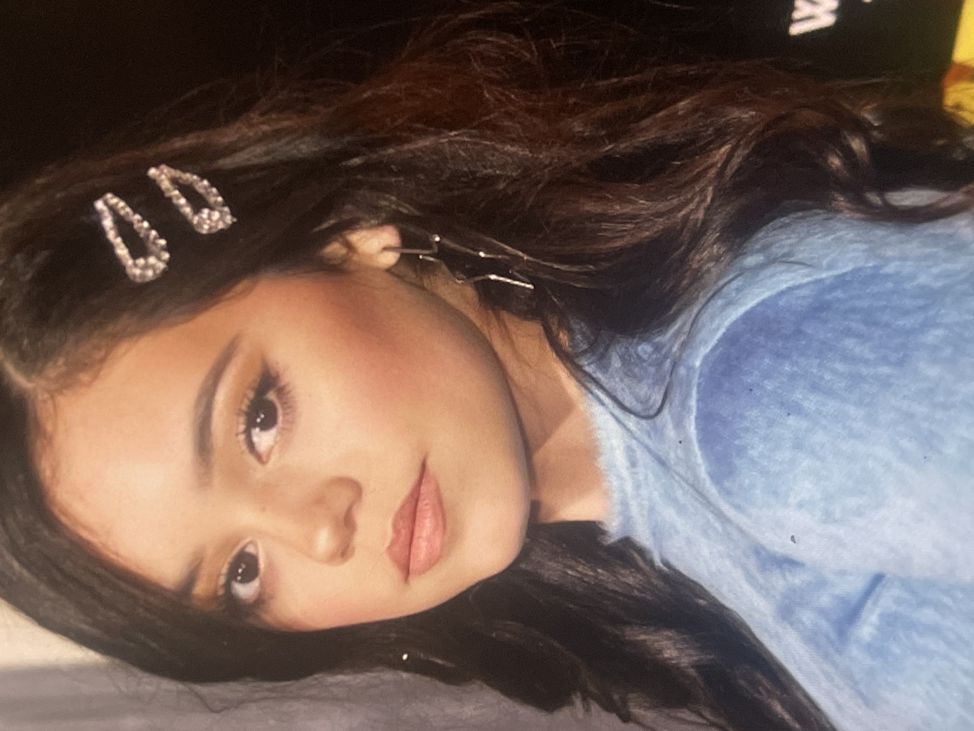 A close-up portrait, oriented diagonally, features a young woman with long, dark, wavy hair. She is looking directly at the camera with a neutral to serene expression. Her face is well-lit, highlighting her features, including defined eyebrows, dark eyes, and light pink or nude lipstick. A flush of blush is visible on her cheeks.
Her hair is adorned with two sparkling, teardrop-shaped rhinestone clips above her left temple. On the right side of her head, near her ear, a delicate, metallic ear cuff or earring with thin, wire-like extensions is visible, partially intertwined with her hair. She is wearing a light blue, possibly fuzzy or textured, long-sleeved top or sweater.
The background is dark and indistinct, suggesting an indoor setting. A faint white letter "W" can be discerned in the upper right portion of the background. The overall lighting is soft, casting a gentle glow on her face. The image, provided with a location context of Poltava, Ukraine, captures a posed moment with a focus on personal adornment and expression.