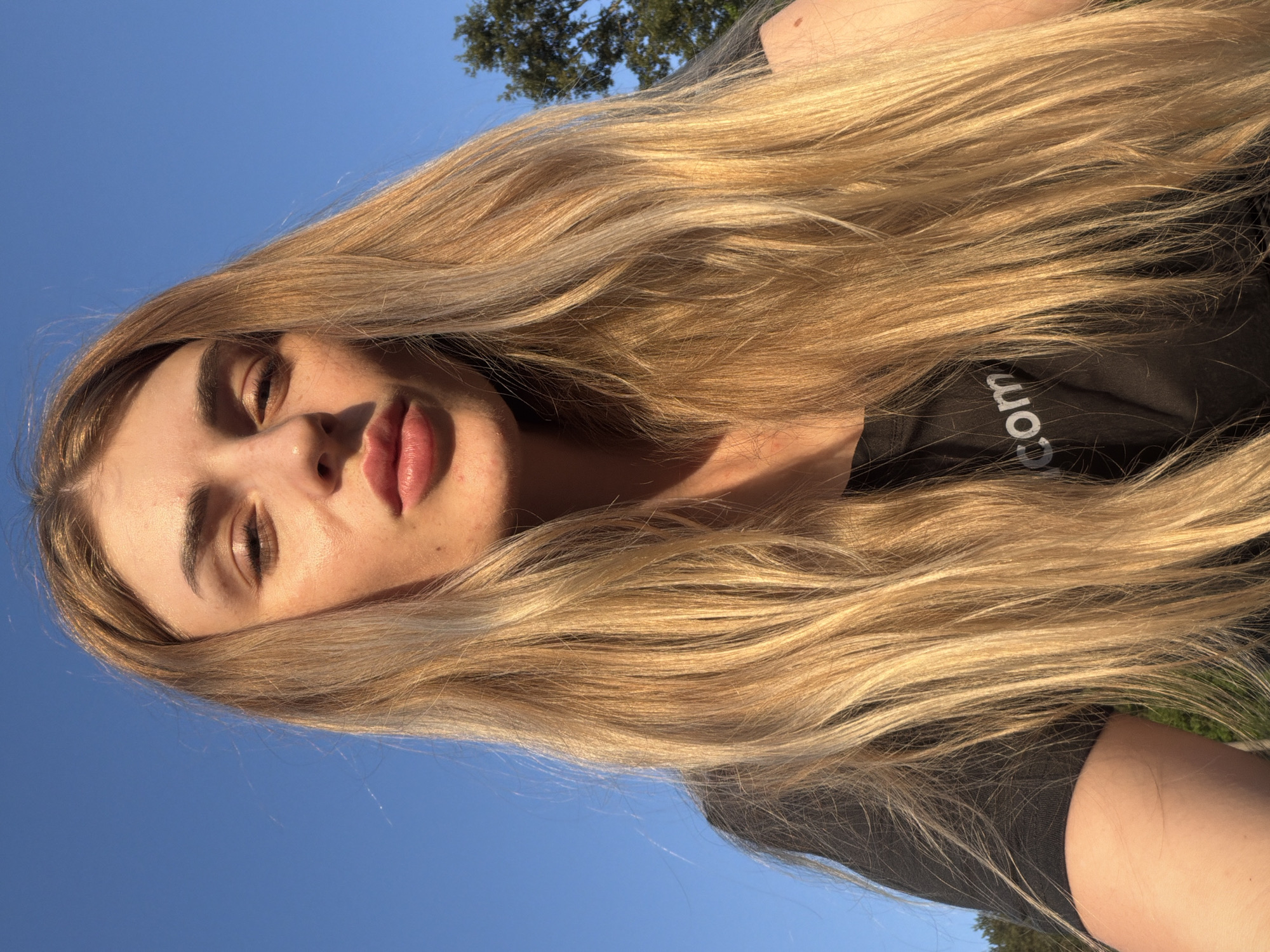 An outdoor, close-up, selfie-style shot shows a young woman with long, blonde hair against a bright blue sky. She is wearing a black t-shirt with the partial word "com" in white lettering on it. The sunlight casts a warm glow on her face, highlighting her features and creating shadows. Her lips are pursed slightly. In the background, the upper branches of a tree are visible on the right side of the frame. The scene appears to be in an unspecified urban or suburban outdoor location.