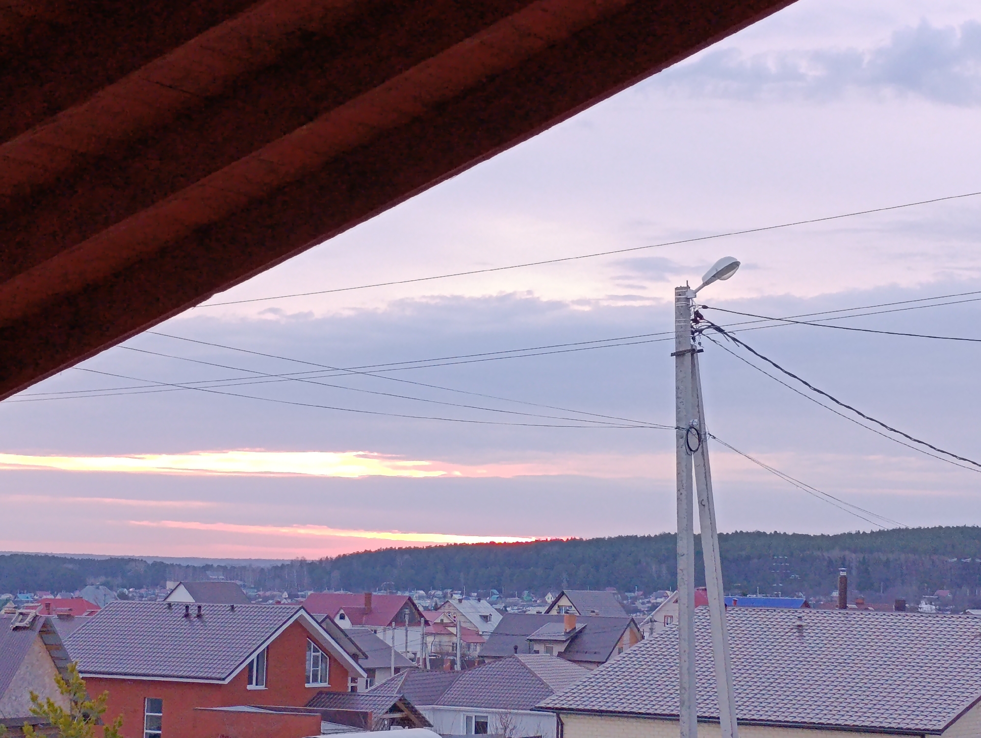 A scenic view from an elevated position overlooks a residential area in the city of Chaikovsky, Russia, under a sky painted with the soft hues of dusk or dawn. A section of a corrugated metal roof in the foreground partially frames the vista. Below, houses of varying sizes and designs are densely packed together, their roofs mainly tiled in shades of brown and grey. Interspersed between the dwellings is a forest line, marking the horizon.
A utility pole with a street light stands prominently in the middle ground, with electrical wires stretching across the sky. The light isn't turned on. The sky is dominated by clouds tinged with pink and purple, suggesting either sunrise or sunset. The overall impression is one of a quiet, settled suburban area.