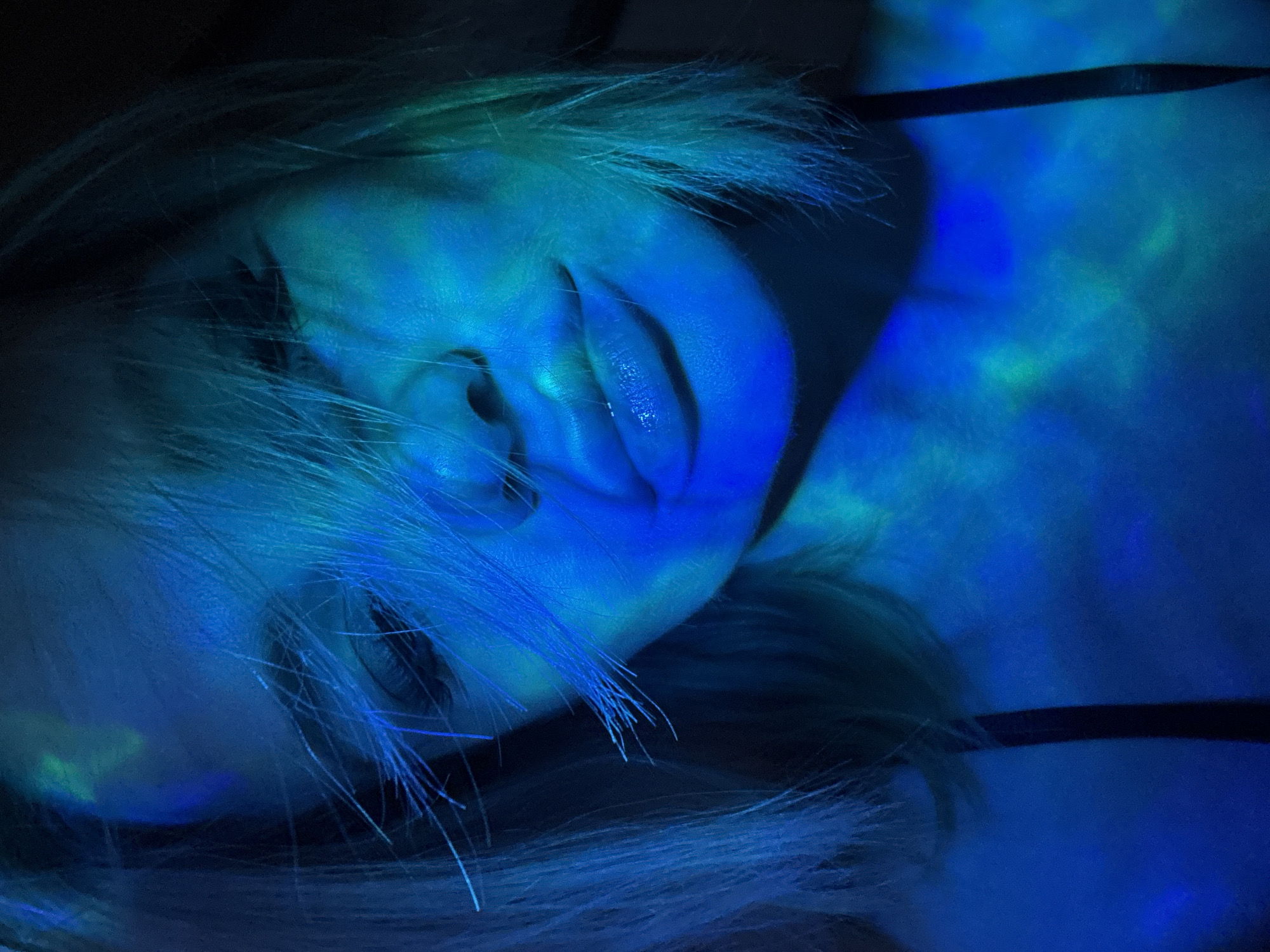 A close-up, low-angle image features a person lying down, illuminated by blue light with hints of green. The person has fair skin and light-colored hair, some strands of which fall across their face. Their eyes are mostly closed, and their mouth is slightly open. A thin black strap, possibly a garment, is visible across their upper chest/shoulder area. The background is dark, adding to the dramatic lighting effect. The location is not identifiable from this image.