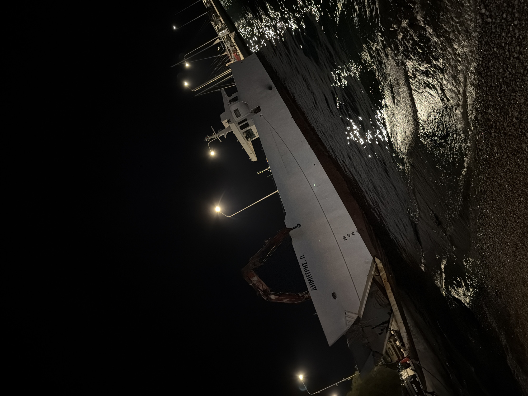 At night, a white ship named "DIMITRIS P." is partially grounded on a pebble shore in the Municipal Unit of Alyzia, Greece. The ship is illuminated by streetlights and reflections from the water. A crane is extended from the ship. The water is dark with shimmering light reflecting on its surface. In the background, there are more lights from streetlamps and parked cars on a paved area.