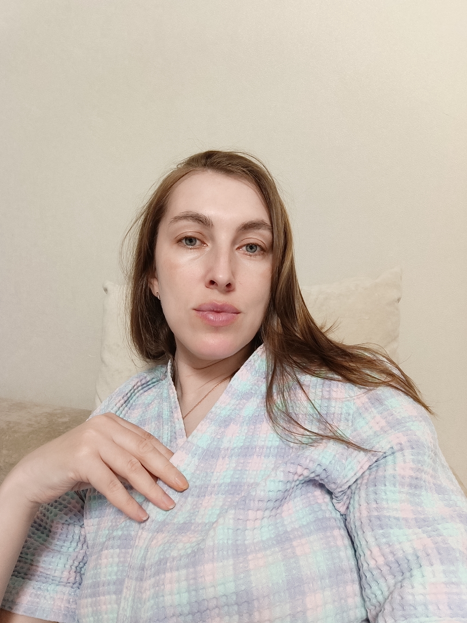 A woman with light skin, blue eyes, and brown hair is in a seated position, looking directly at the camera. She's wearing a pastel plaid robe. Her right hand is resting on her chest. She is sitting on a beige surface with a light-colored pillow visible behind her. The background is a plain, light-colored wall. There is no information to confirm the location in Chaikovsky, Russia.