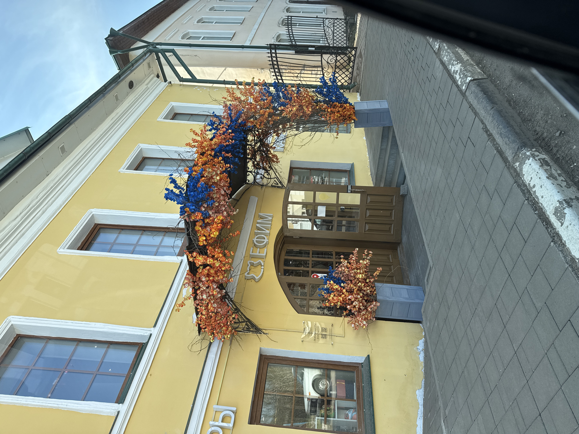 A ground-level shot of a building exterior in Ново-Никольское, Russia. The building is yellow with brown trim around the windows and doors. Blue and orange floral decorations surround the entrance. The name "ЕФИМ" is displayed above the entrance. A stone sidewalk runs alongside the building.
