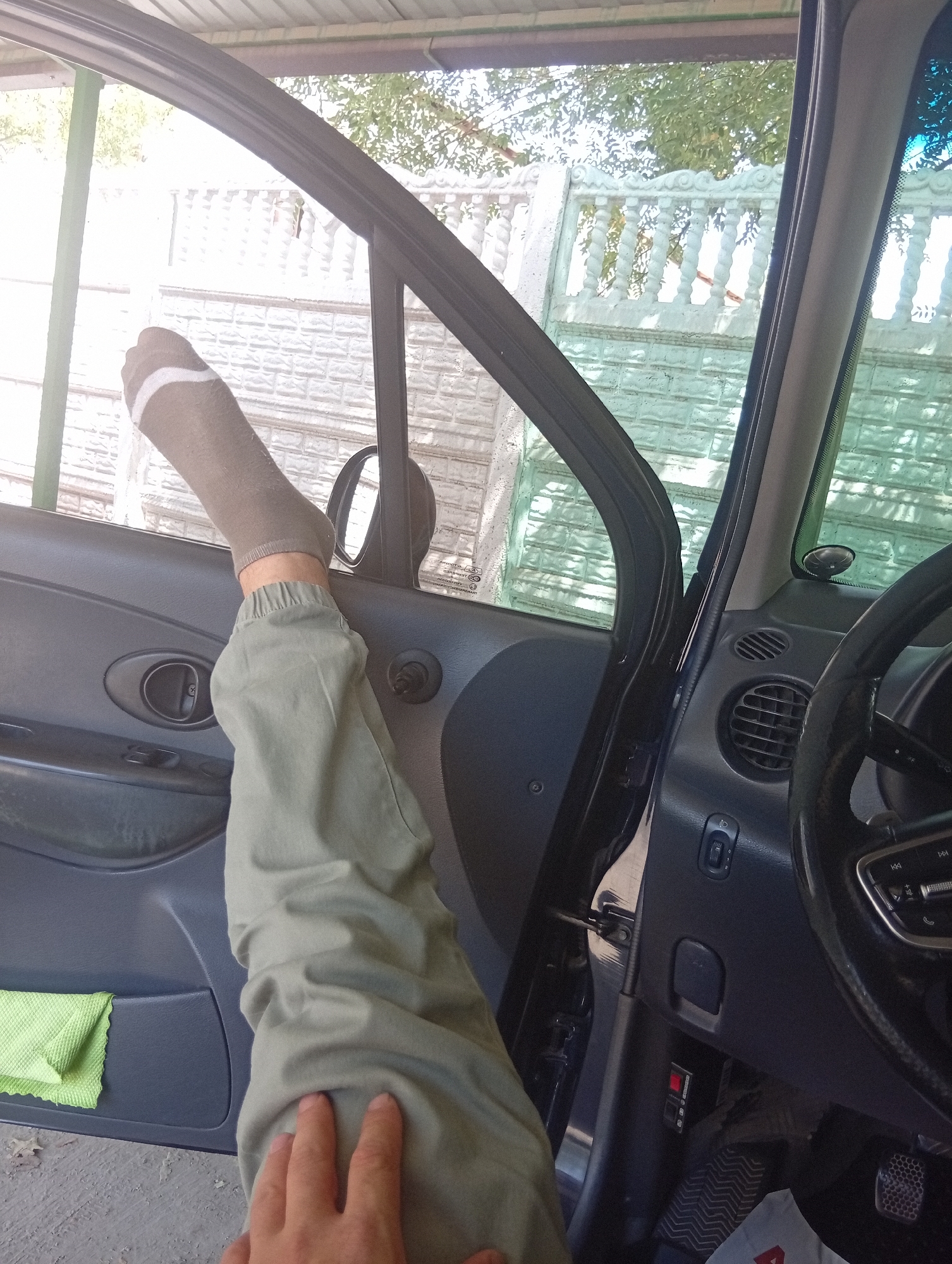 The image shows a person's leg, clad in a grey sock and light green pants, extended out of the open driver's side door of a car. The car's interior, including the door panel and steering wheel, is visible. Outside the car, a white ornate fence and a textured wall are visible, set against a backdrop of trees and a sunny sky. This scene is likely located in Tashkent, Uzbekistan, characterized by the typical architecture and vegetation found in the region.