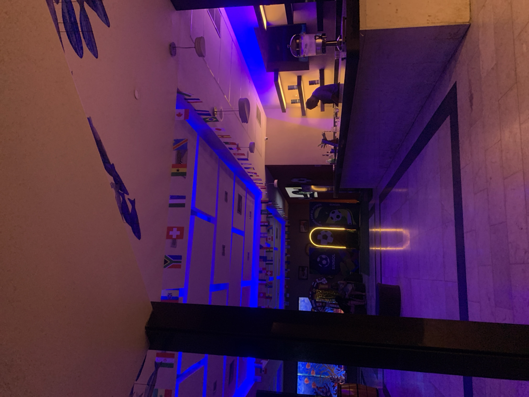 The image depicts the interior of a bar or establishment in El Tirano, Venezuela. The scene is illuminated by predominantly purple and blue lighting, casting a vibrant and somewhat atmospheric glow. In the foreground, a dark structural pillar occupies the left side of the frame. The floor is tiled with a light-colored material, accented by darker grout lines, creating a patterned effect.
The ceiling features a drop ceiling with some panels illuminated by blue LED strip lighting. Along the upper edges of the walls, a string of small national flags is hung, suggesting an international theme or perhaps a connection to a sporting event. Decorative elements, possibly depicting stylized blue fish or aquatic creatures, are visible on the sloped sections of the ceiling.
In the middle ground, to the left, is a large aquarium or illuminated display featuring what appear to be orange fish. Further to the right, a bar area is visible. Behind the bar, a person, likely a bartender, is standing and appears to be working. Shelving with some items is present on the back wall behind the bar. In the central area of the establishment, a distinctive illuminated sign with a yellow outline in the shape of a horseshoe or a glowing arch is present, with what appears to be a soccer ball graphic and the word "SUNSOL" visible on banners. Two individuals are seated in the background near the aquarium.
On the right side of the frame, a silver beverage dispenser with a tap is positioned on the bar. The overall environment suggests a recreational or hospitality setting, possibly a sports bar or a themed lounge.