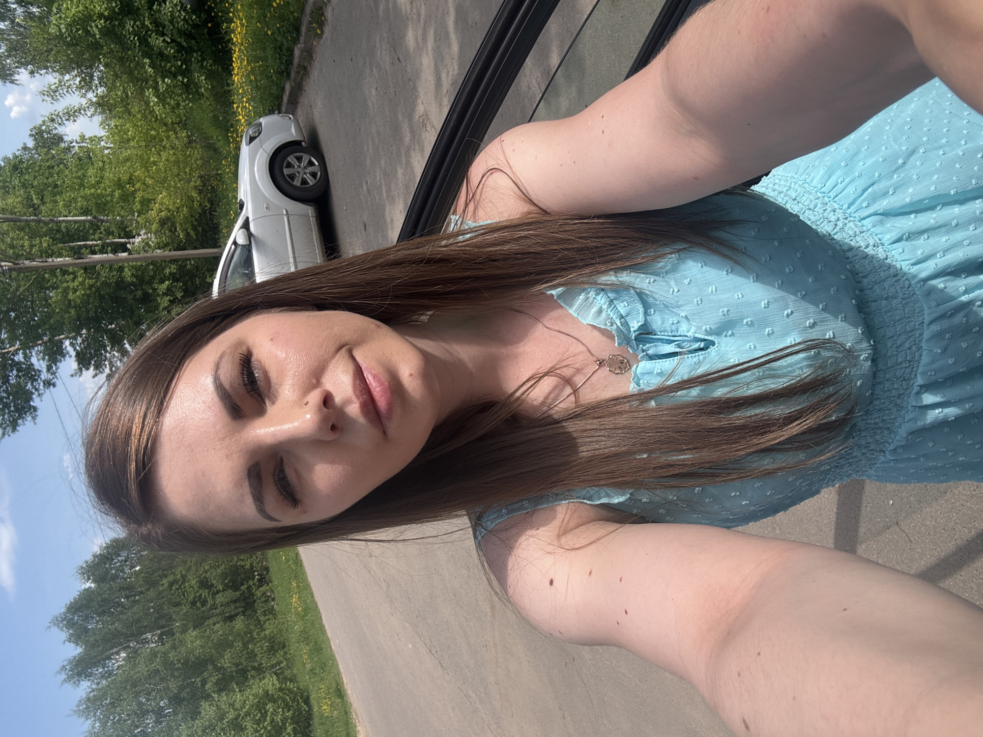 A woman is taking a selfie outdoors. She is wearing a light blue dress and a necklace. A white car is visible in the background, parked on the side of the road. Trees are also visible in the background. The location is potentially Shevlyakovo, Russia, but there is no way to verify it from this image.
