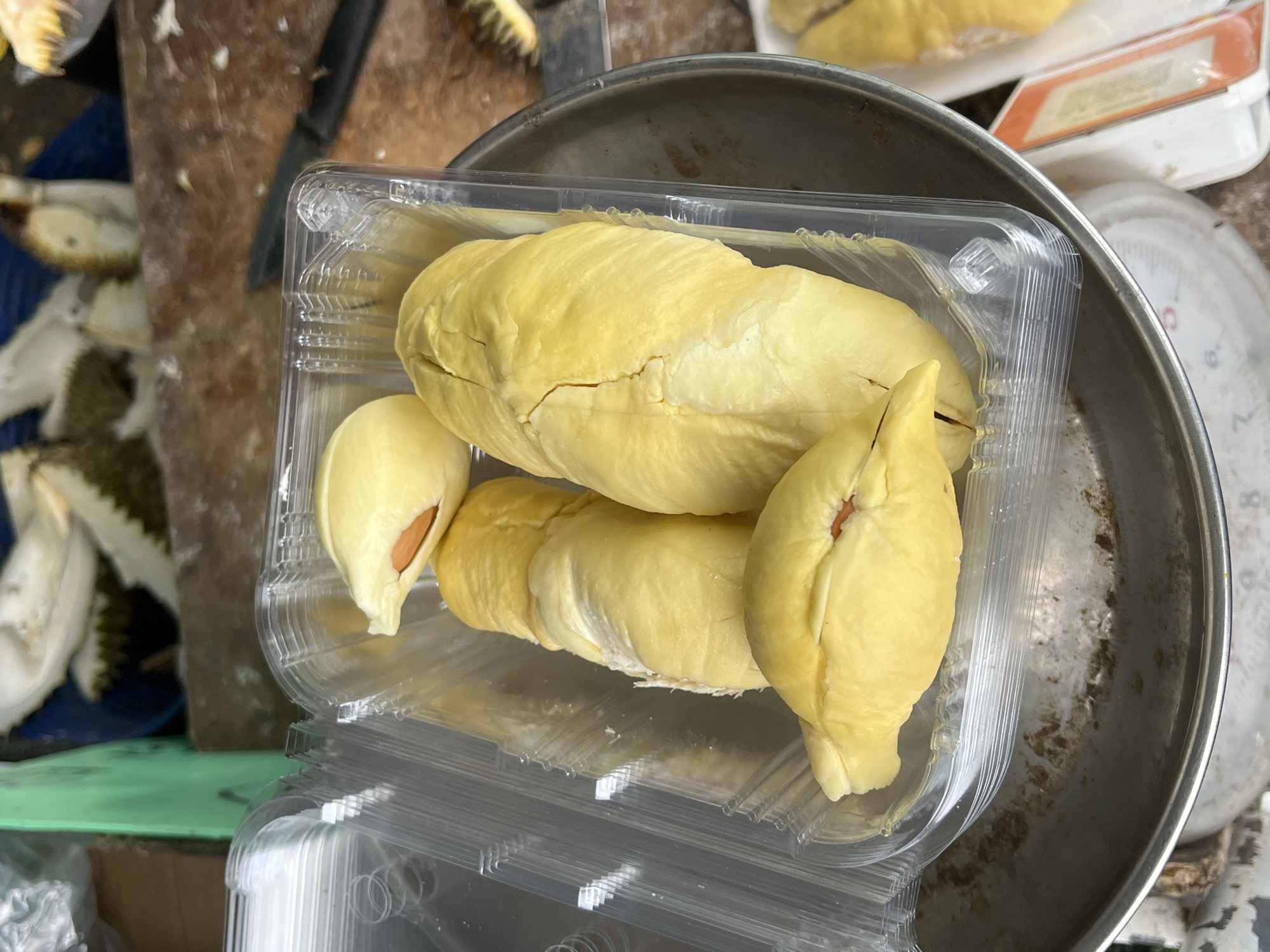 In Rawai, Thailand, a clear plastic container holds four pieces of peeled durian. The durian flesh is a pale yellow, with some pieces showing a prominent orange-brown seed. The container is placed on a round metal weighing scale. In the background, parts of a durian fruit with spiky green skin are visible, along with a green plastic object and a wooden surface.