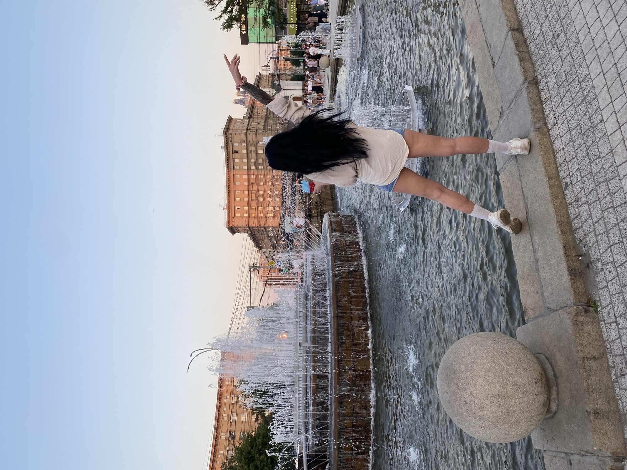 A young woman with long dark hair stands facing away from the camera, holding a blue and red cup, and with her left arm outstretched to the side. She is wearing a loose-fitting cream-colored shirt and denim shorts, white socks, and white sneakers. She is standing on the edge of a large fountain in the city of Novosibirsk, Russia, with water spraying upwards. In the background, there are several multi-story buildings, some with ornate architectural details, and streetlights. The sky is clear and a pale blue, suggesting it is either early morning or late afternoon. The overall scene is urban and depicts a public space with a fountain and surrounding buildings. The woman appears to be enjoying the outdoor environment.