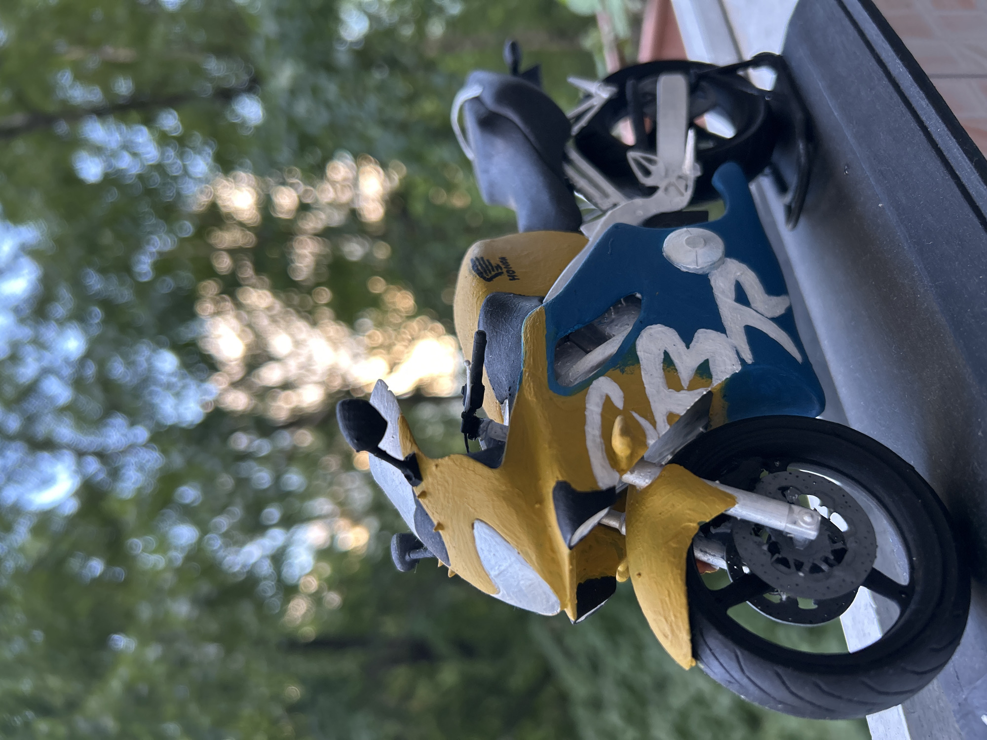 A close-up shot captures a miniature motorcycle model, prominently featured in the foreground. The model is primarily yellow and blue, with black and white accents. The front fairing and lower side panels are yellow, while the fuel tank area is blue with stylized white lettering, possibly depicting "MR" or a similar design. A small, black "HONDA" logo is visible on a yellow section near the handlebars, alongside a claw-like graphic. A miniature black helmet rests behind the handlebars. The front wheel, with its disc brake, is clearly visible in the lower right, displaying intricate spoke details and a textured tire.
The background is heavily blurred, showcasing green foliage and numerous bright, circular bokeh lights, indicating natural light filtering through trees. This suggests the photo was taken outdoors during the daytime under sunny conditions, with a shallow depth of field. The overall image is rotated 90 degrees clockwise, giving the motorcycle a vertical appearance. The model appears to be made of plastic or a similar painted material.