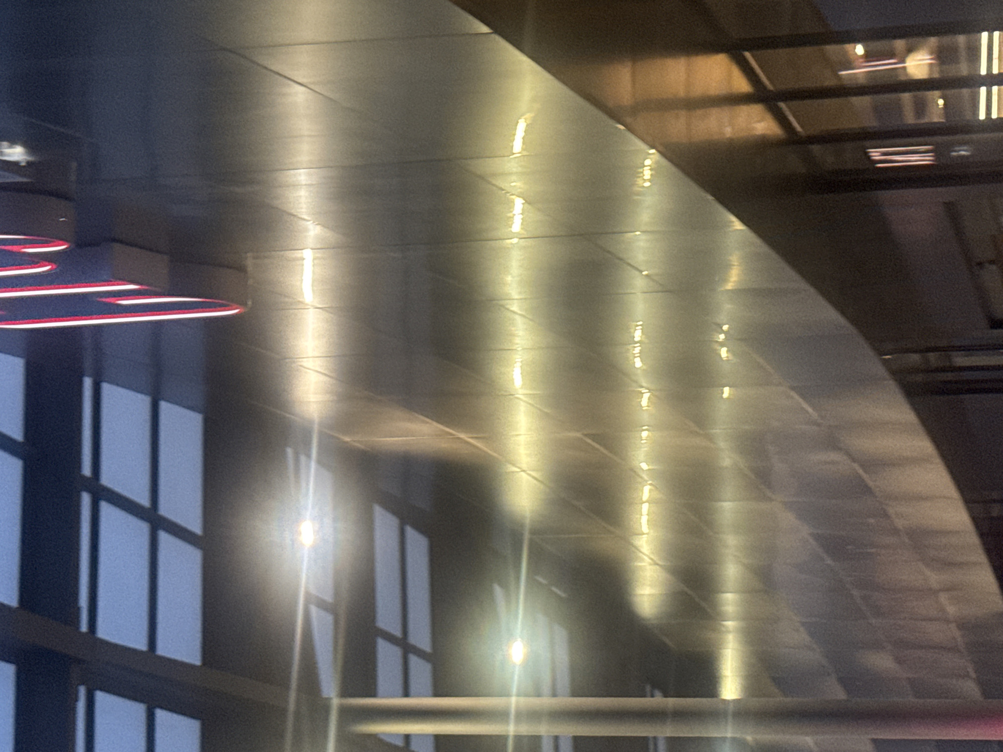 The image shows the curved ceiling of a modern building, likely an airport or transportation hub, in Puebla, Mexico. The ceiling is constructed of dark, reflective metal panels, creating a grid-like pattern. Artificial lighting is reflected in the panels, appearing as bright, vertical streaks. Below the ceiling, a window wall with multiple panes of glass is visible, showing the exterior light and sky. In the left corner, a portion of a red and white neon or LED sign is seen. The overall impression is a modern, industrial space with diffused and reflected light.