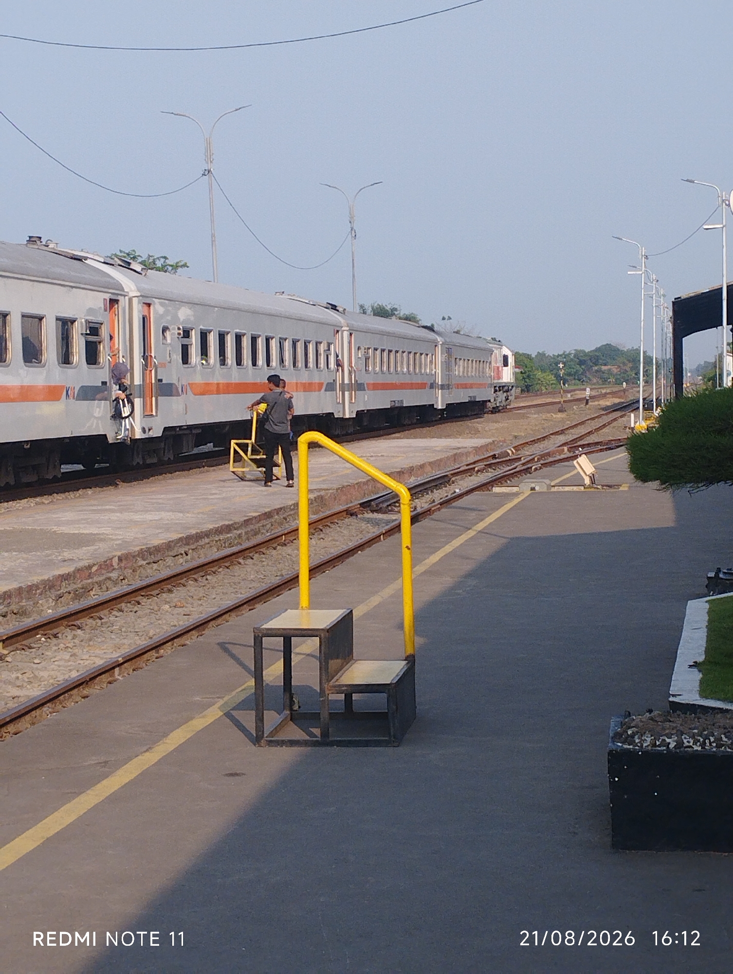 The image depicts a train station platform in Karawang, Indonesia, on August 21, 2026, at 16:12. A silver passenger train with orange stripes is parked at the platform. Two individuals are visible on the platform near the train. One person, wearing a dark shirt and pants, is standing on the platform next to a yellow metal step stool. Another person, wearing a dark shirt and lighter pants, is seen near the entrance of one of the train carriages. The platform is paved with concrete and bordered by railroad tracks. A bright yellow railing and a black metal structure with a seat are also present on the platform. In the background, there are streetlights, trees, and more railroad tracks extending into the distance. The sky is clear and pale blue, suggesting daylight.