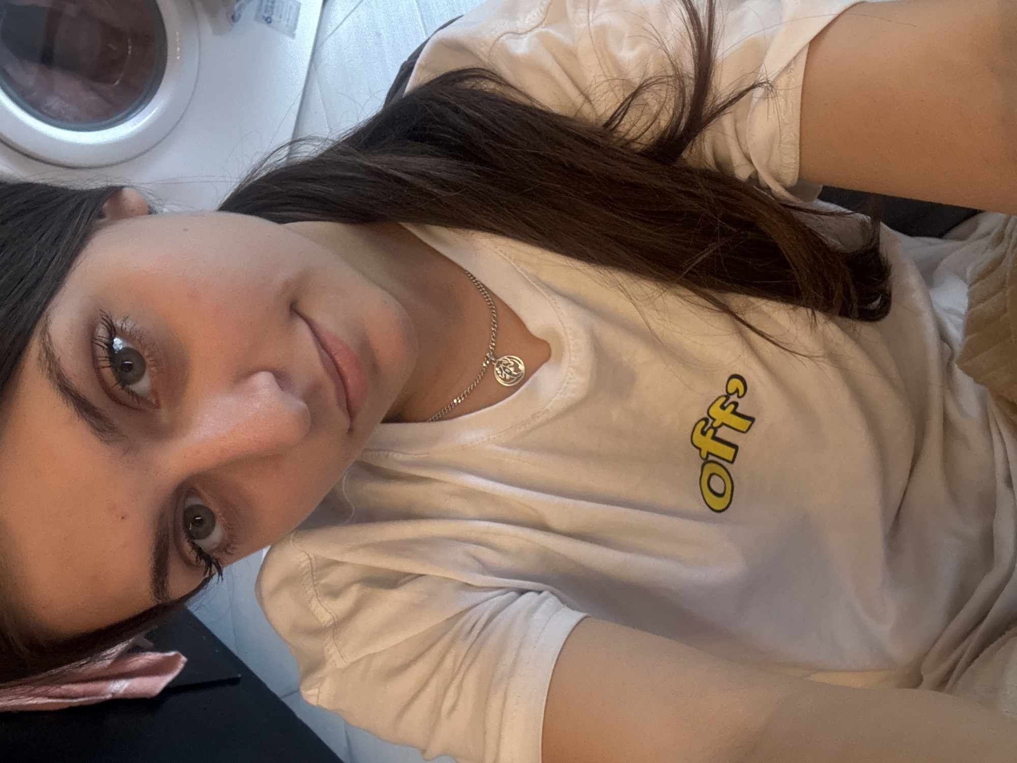 A selfie shows a young woman with brown hair and green eyes wearing a white t-shirt with the word "off" printed on it in yellow. She is also wearing a silver necklace. Behind her is a white washing machine and some tiled flooring. No further details about the location in Pomorie, Bulgaria are discernable.