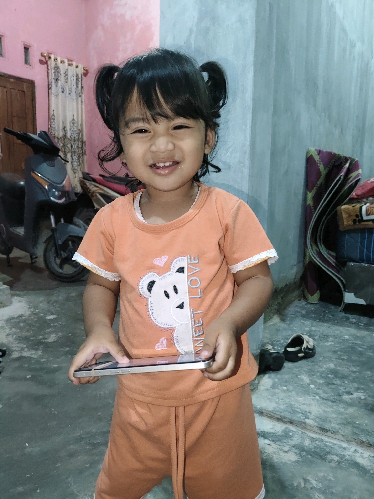 A young girl with dark hair styled in pigtails stands indoors, holding a smartphone horizontally in her hands. She is wearing an orange t-shirt with a bear graphic and the text "SWEET LOVE" and matching orange shorts. The background includes a pink wall with cream-colored curtains, a dark-colored scooter, and a light blue concrete wall with some stacked items. The floor appears to be a gray concrete surface. The girl is smiling and looking at the camera. This scene appears to be inside a residence in Fajar Baru, Indonesia, possibly a living room or a covered outdoor area adjacent to the main house.