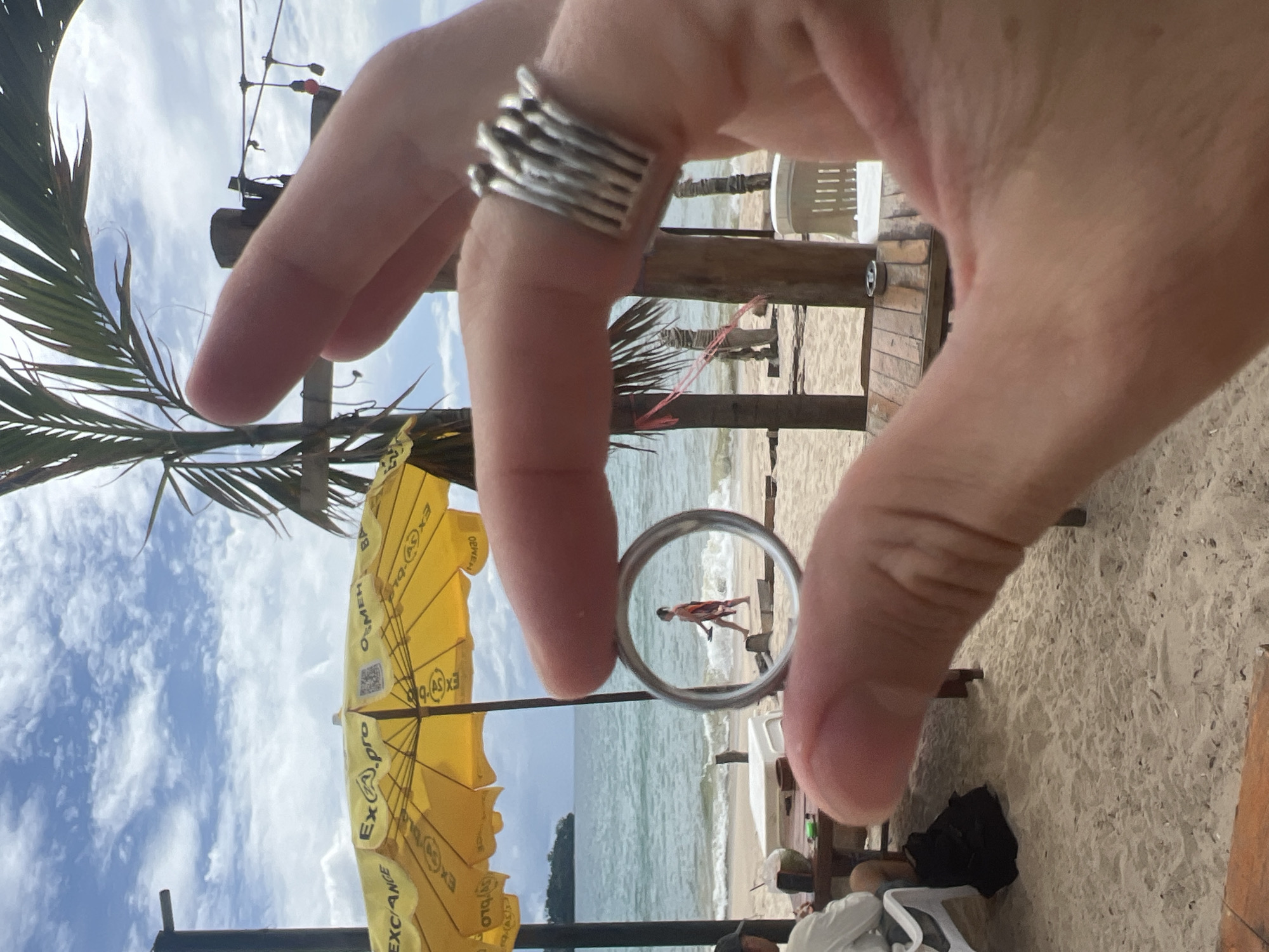 A person's hands are in the foreground of the image, holding a ring up to frame a scene on a beach in Choeng Thale, Thailand. The ring is silver and has a stacked, linear design. Through the ring, a person in a colorful sarong is walking on the sand, carrying a flip-flop. In the background, a yellow umbrella with "EXCHANGE" printed on it provides shade for a beachside area. Palm trees and a cloudy blue sky are visible above. The beach is sandy, with the ocean lapping at the shore. A few wooden tables and chairs are set up on the sand.