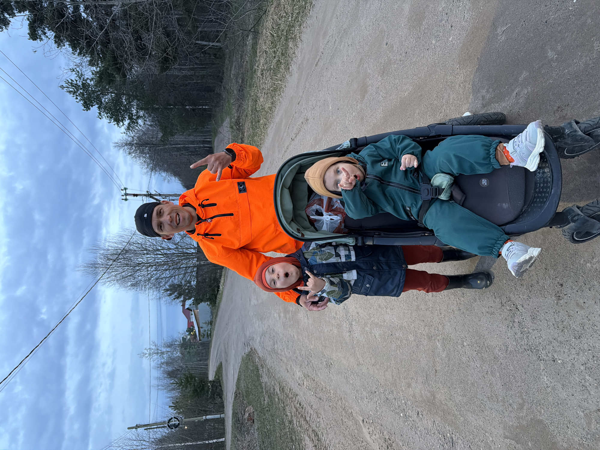 The image, oriented in portrait, captures a man and two young children outdoors on a dirt road, likely in Chaikovsky, Russia, under an overcast sky.
The man, positioned on the left, wears a striking bright orange jacket with a black patch on the left chest displaying a white logo (possibly "SWEEP"), a black beanie, and dark trousers. He is smiling broadly and making a "shaka" or "rock-on" hand gesture with his right hand.
To his right, a child is seated in a dark-colored stroller or wagon. This child wears a green full-body jumpsuit with a white patch on the left arm featuring a black logo (possibly two stars), a light brown hooded top, and light grey sneakers with orange socks. This child is also smiling and making a similar "rock-on" hand gesture with their left hand. A red and white plastic bag is visible inside the stroller.
Standing between the man and the stroller, slightly in front, is another child. They are dressed in a dark blue patterned jacket, red pants, a red hood, and black boots. This child is looking towards the camera with a playful expression.
The setting is a rural or semi-rural dirt road flanked by a forest of mostly bare trees, suggesting late winter or early spring. Power lines stretch across the sky, supported by a utility pole visible on the left. A building with a red roof can be seen in the distance on the right. The overall atmosphere is lighthearted and casual, with all subjects appearing engaged with the camera.