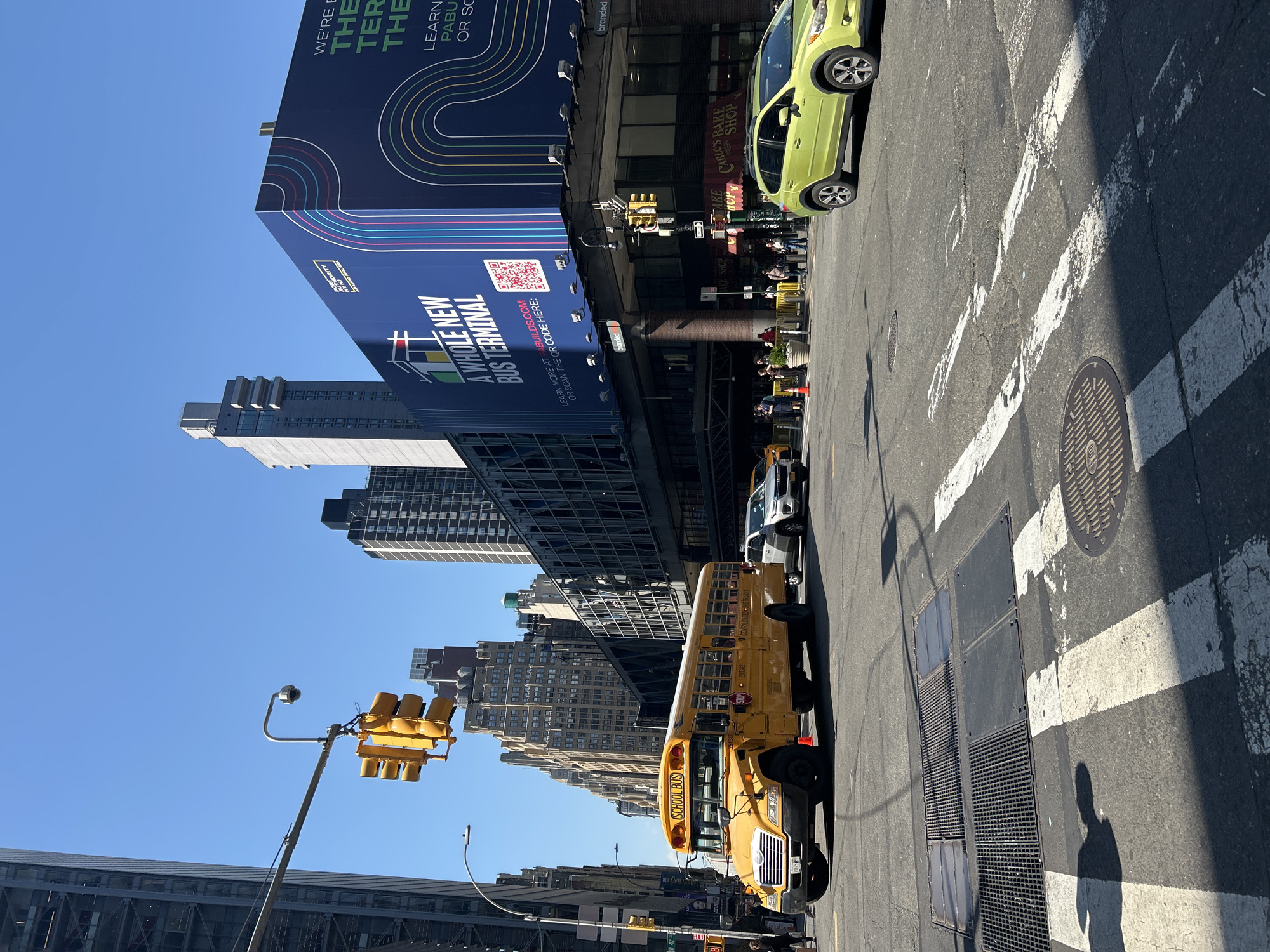 This is a street scene in New York City, United States of America. A yellow school bus is parked on the side of the road. A light green car is also visible. Traffic lights are suspended overhead. A large advertisement for the Port Authority bus terminal is displayed on a building. The city skyline is visible in the background, including a mix of modern and older buildings. A shadow of a person can be seen on the street. There are crosswalk markings on the road. A manhole cover is visible on the road.