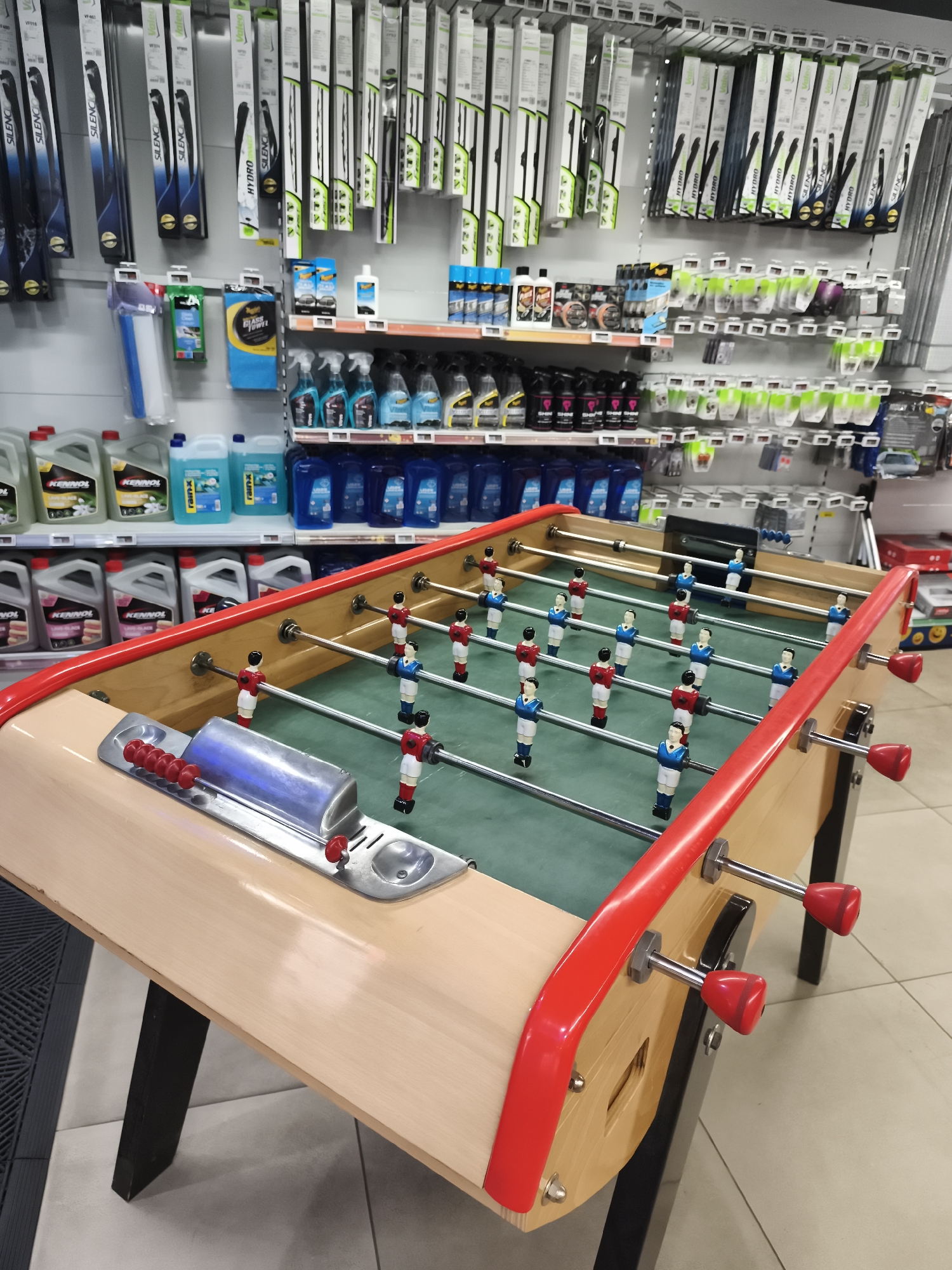 The image depicts a foosball table positioned in the foreground of a retail store. The table, made of wood with red accents, features green felt playing surface and rows of player figures in red and blue uniforms. Behind the foosball table, shelves are stocked with various automotive products, including windshield wipers, car cleaning supplies, and oils. The store environment suggests it is an automotive or general retail establishment. The image is taken at a low angle, looking slightly upwards towards the shelves. The lighting appears to be artificial, typical of an indoor retail space. The scene is static, with no visible people or ongoing activity. The location is a retail store in Saint-Denis, France.