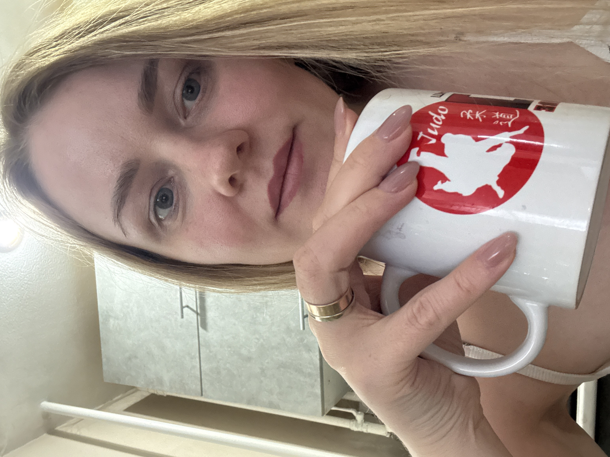 A blonde woman with fair skin holds a white mug with a red circle containing a white silhouette of two figures engaged in a judo stance and the word "Judo" written in English, as well as three Japanese characters, visible against the background of what appears to be a bathroom in Chaikovsky, Russia. The woman's face is angled towards the camera, and her lips have a pinkish hue. She is wearing a gold ring on one of her fingers. The background includes a light grey cabinet, a white pipe, and a partially visible white garment. The scene appears to be indoors, possibly in a private residence.